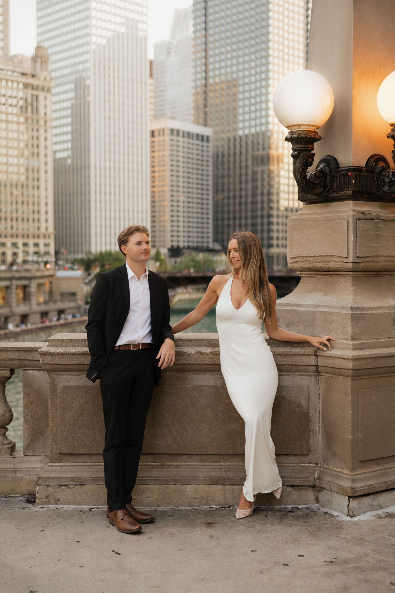Couple posing on a Chicago bridge, man in black suit, woman in white dress, city skyline backdrop.