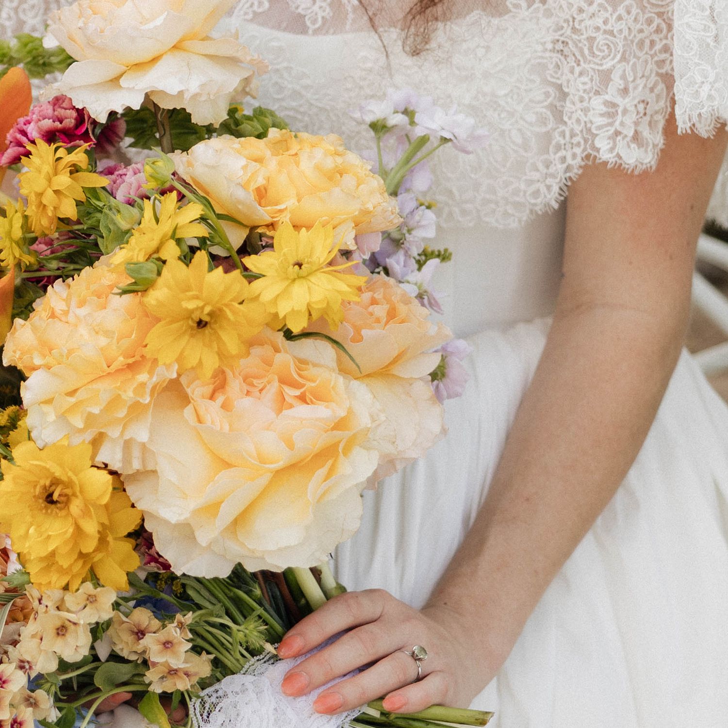 Bride in lace wedding dress holding a vibrant yellow and peach floral bouquet with roses and daisies.