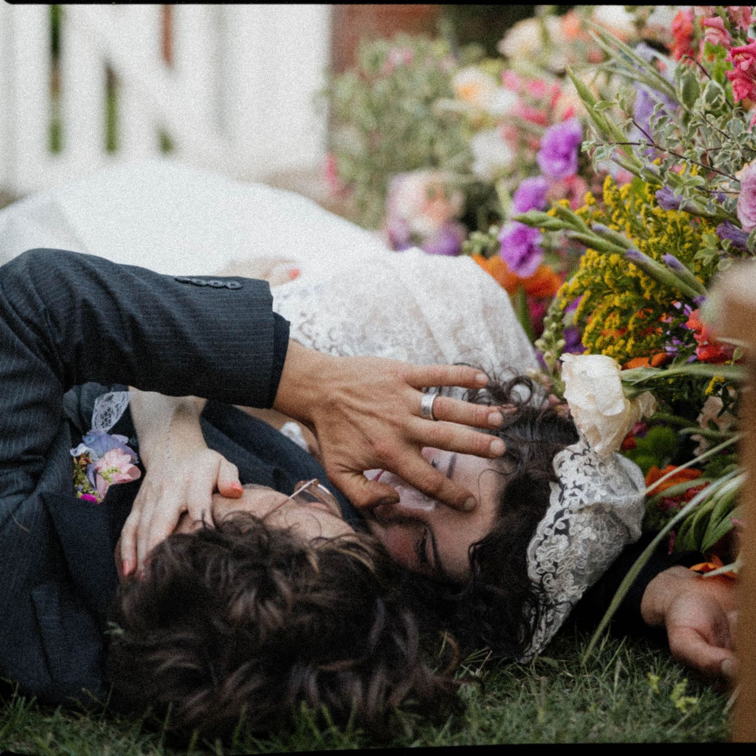 A bride and groom share a romantic kiss while lying on green grass beside a vibrant colorful floral bouquet.