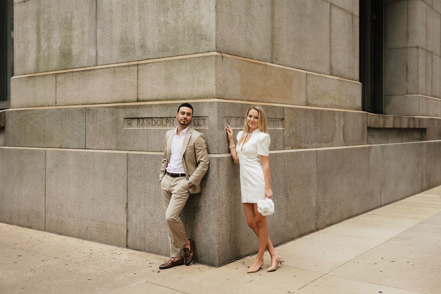 Couple posing against a stone building, man in tan suit and woman in white dress, urban setting.