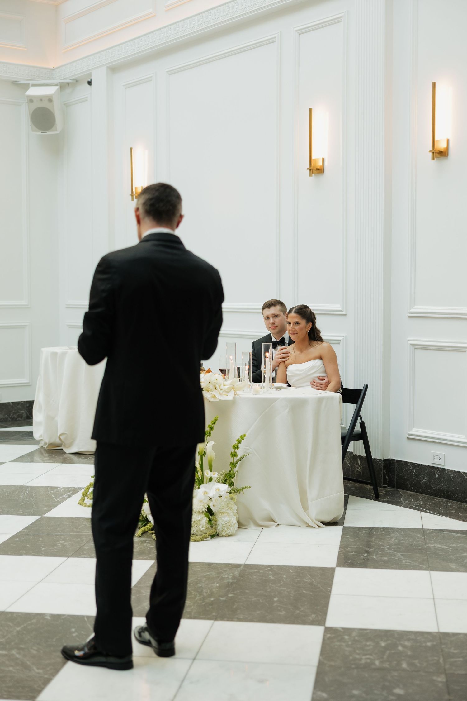 Groom and bride sit at sweetheart table listening to a wedding speech in a white elegant ballroom with gold sconces.