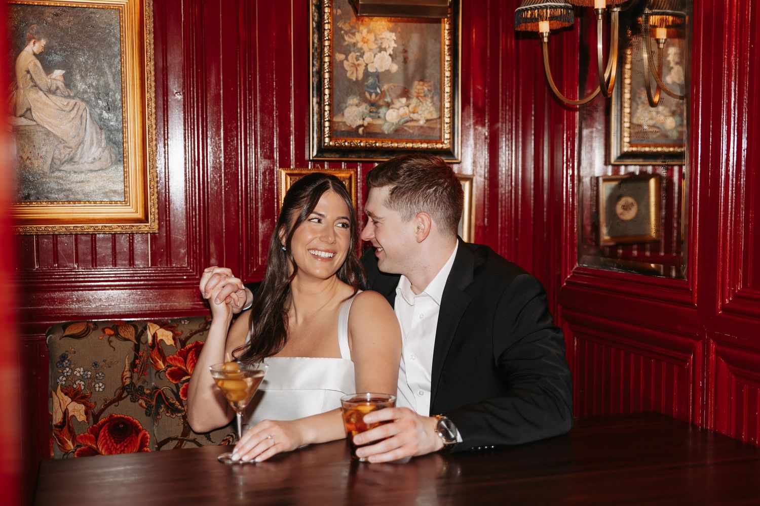 People enjoying drinks and conversation in an elegant red-walled bar with vintage artwork and warm lighting.