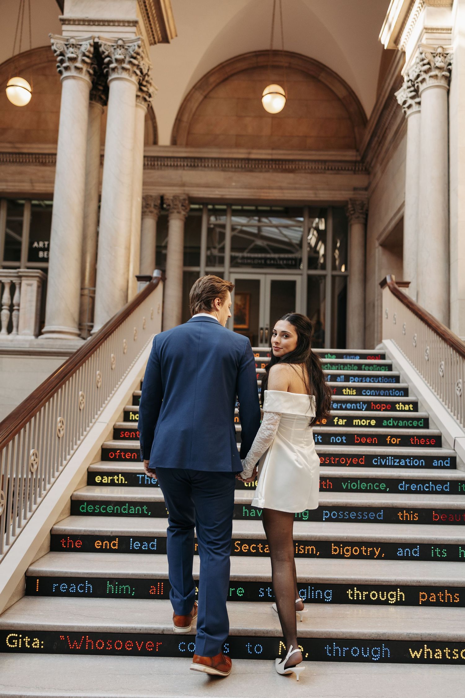 Couple walking up ornate stairs with colorful text written on the risers in an elegant building.