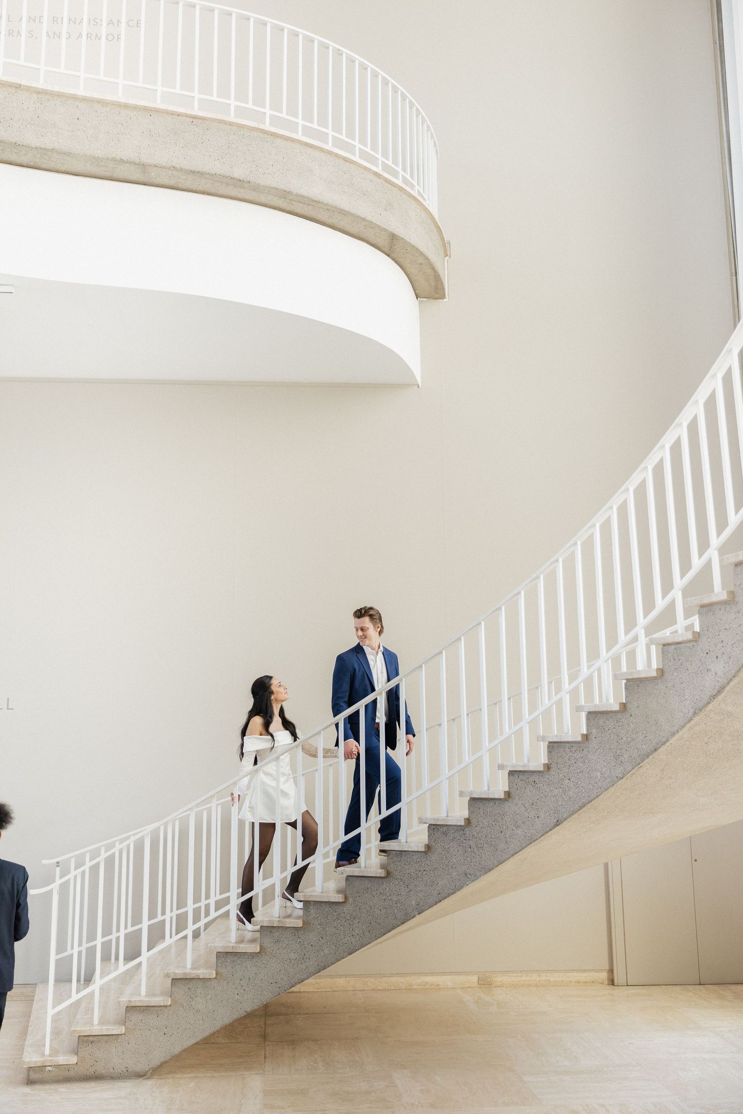 A couple ascends a sweeping white spiral staircase in an elegant architectural setting.