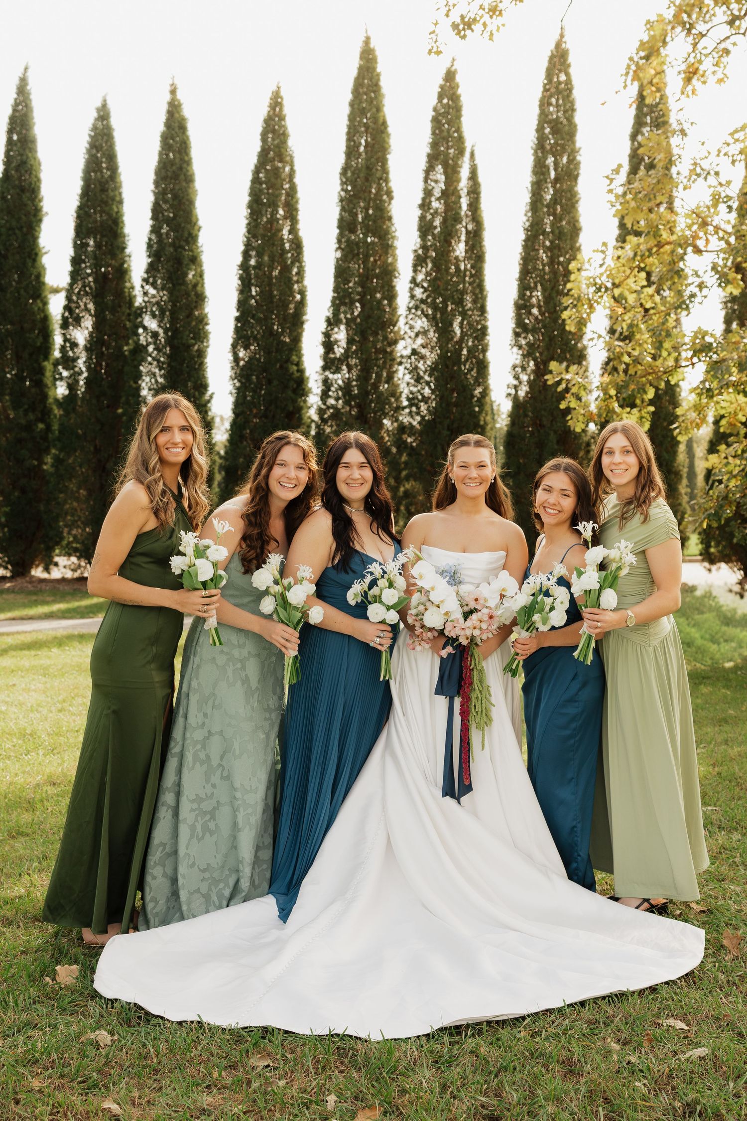 Bride in a white gown surrounded by five bridesmaids in mixed green dresses holding white bouquets before tall cypress trees.