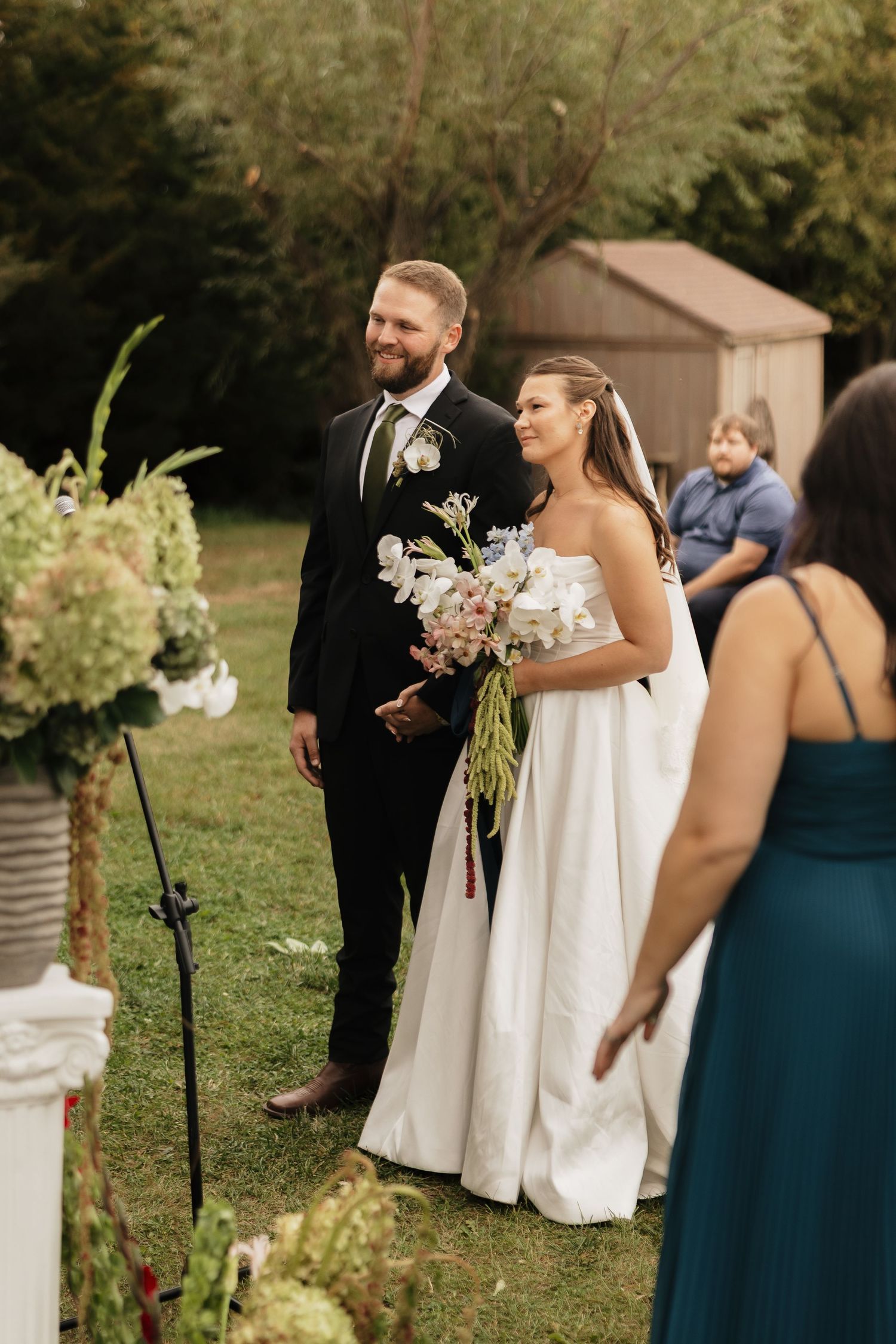 Smiling bride holding a pastel bouquet and groom in a dark suit stand together at their outdoor wedding ceremony.
