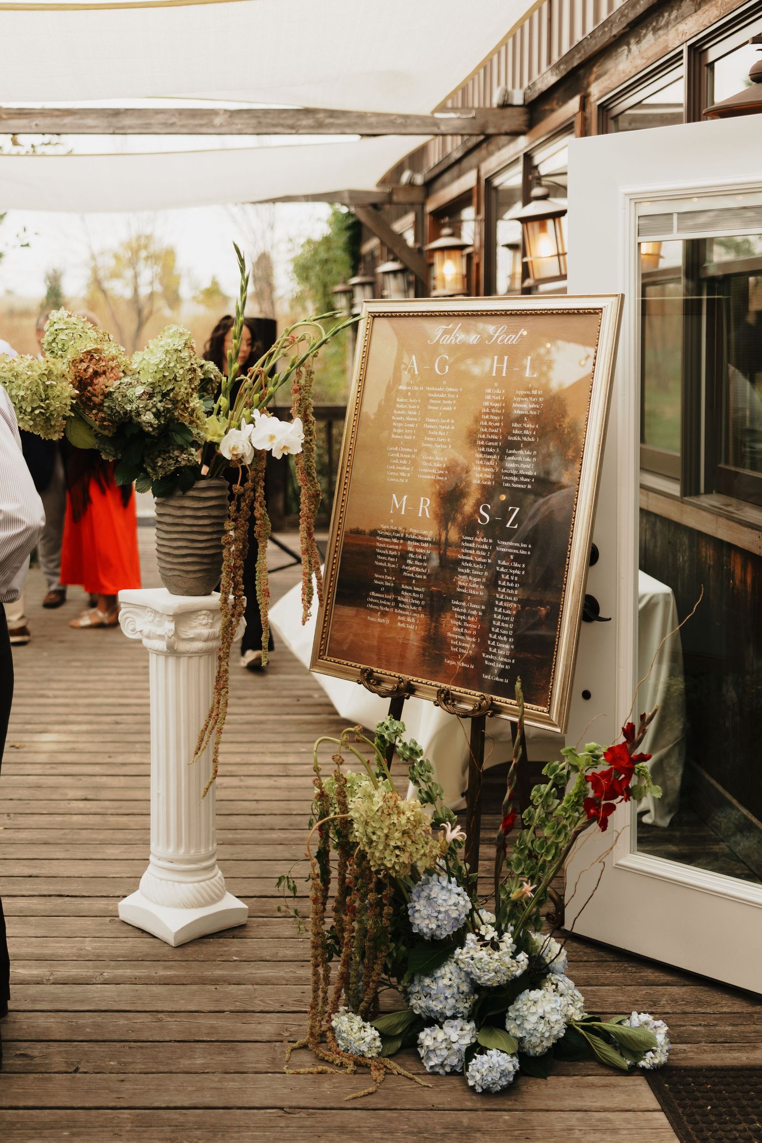 Wedding seating chart on a framed mirror beside blue hydrangeas and a floral arrangement on a white pedestal on a sunlit wooden deck.
