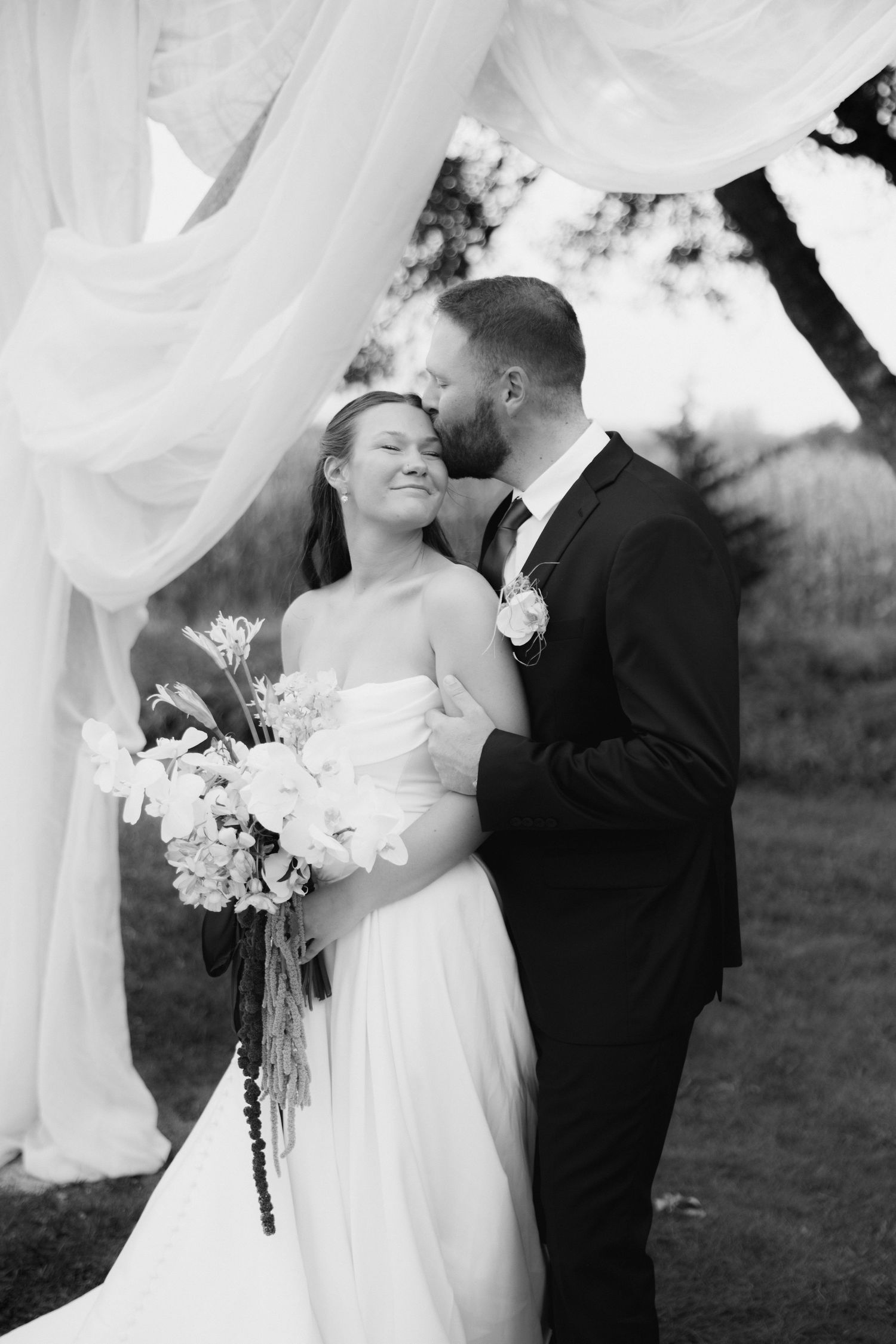 Black and white photo of a groom kissing his smiling bride's cheek under flowing white draping, she holds a wildflower bouquet outdoors.