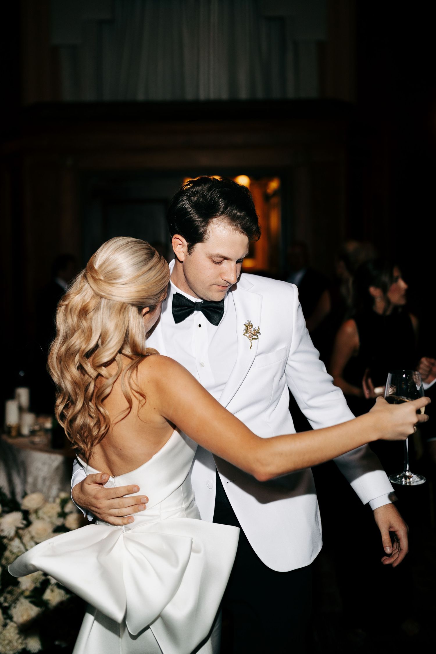 Bride in a white gown with a large back bow and groom in a white dinner jacket and black bow tie share a wedding reception dance.