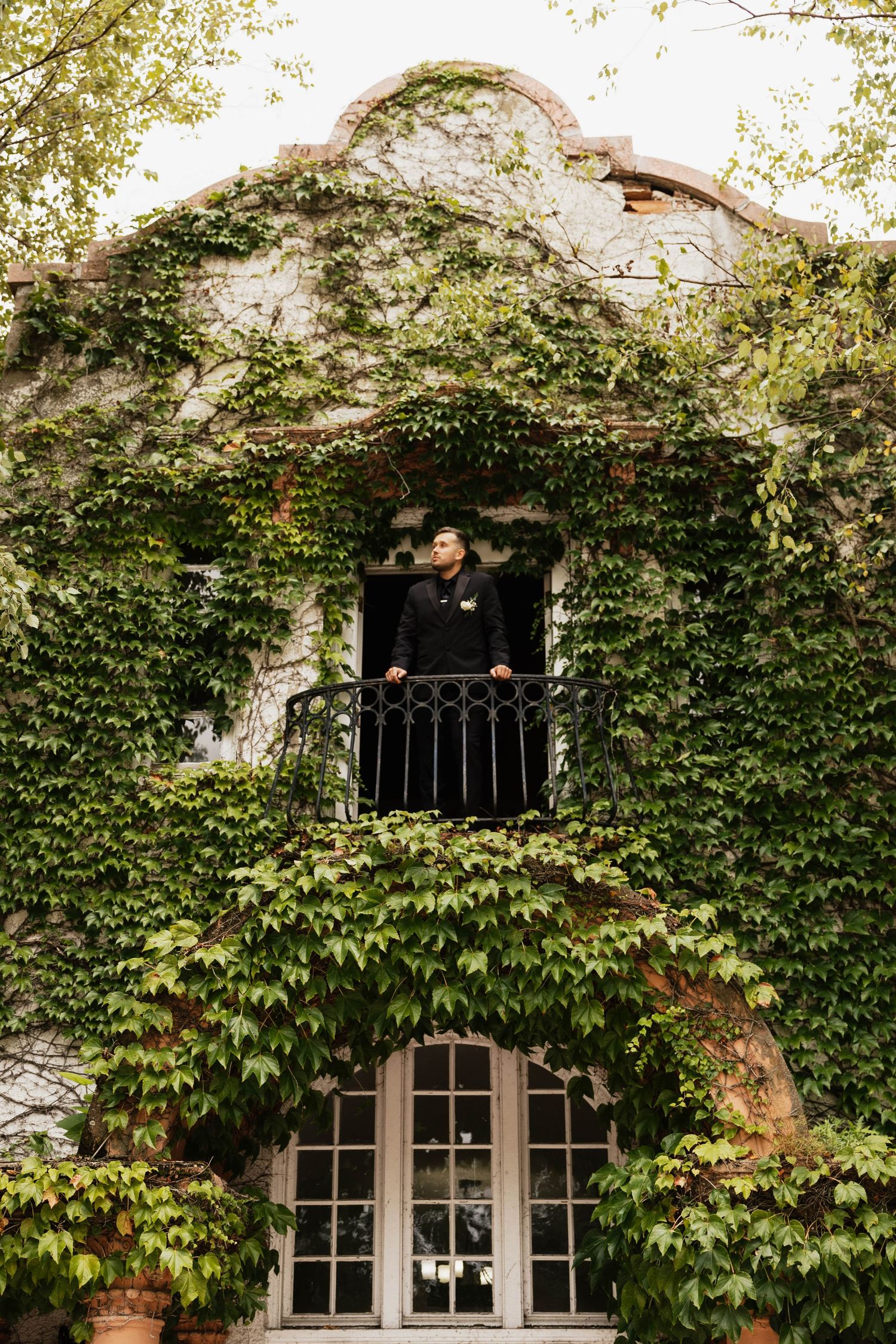 Man in dark clothing stands on a wrought iron balcony of an ivy-covered stucco building with French doors and lush green vines.