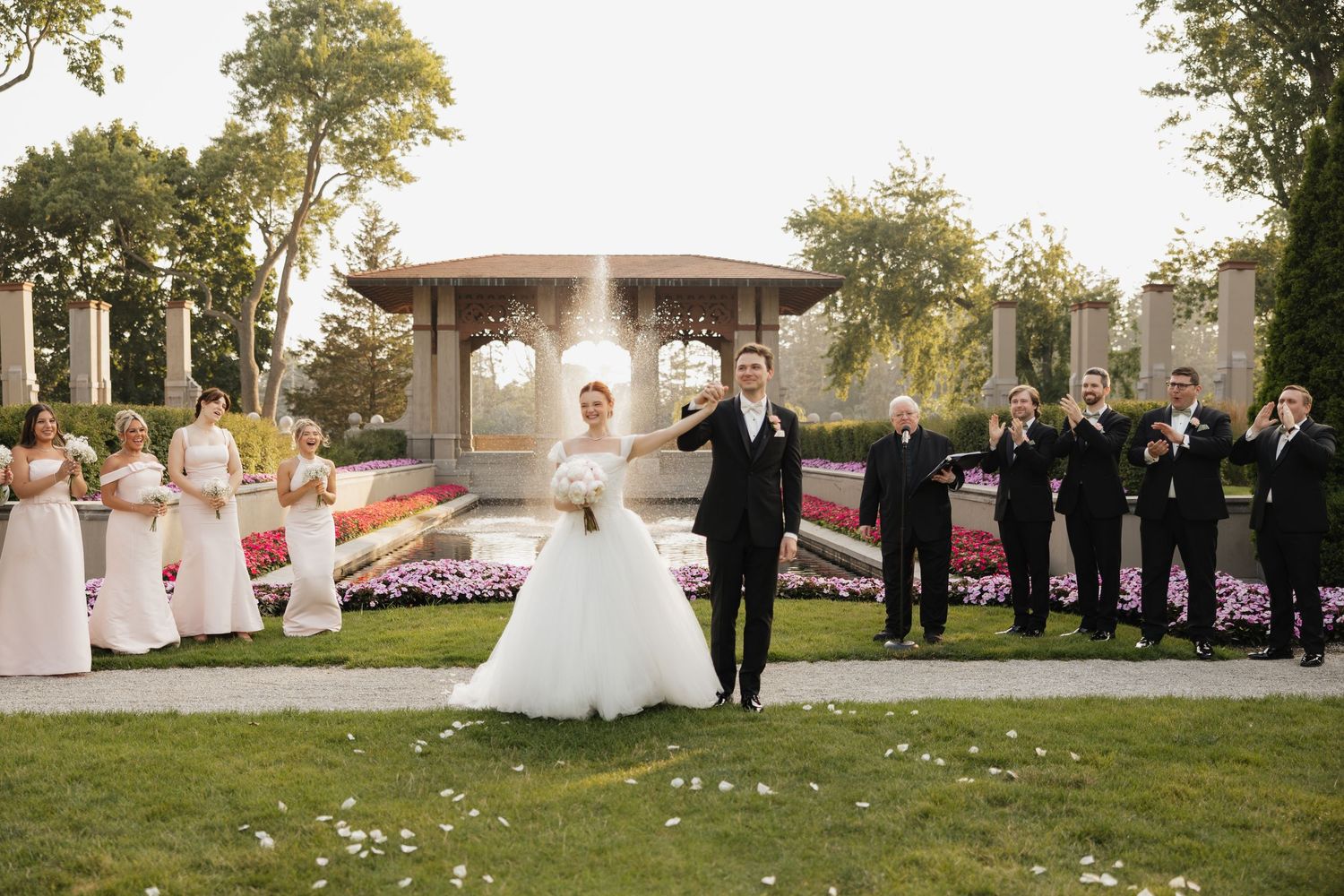 Wedding party poses outdoors near fountain, bride in white gown with groom in tuxedo, bridesmaids and groomsmen celebrating.