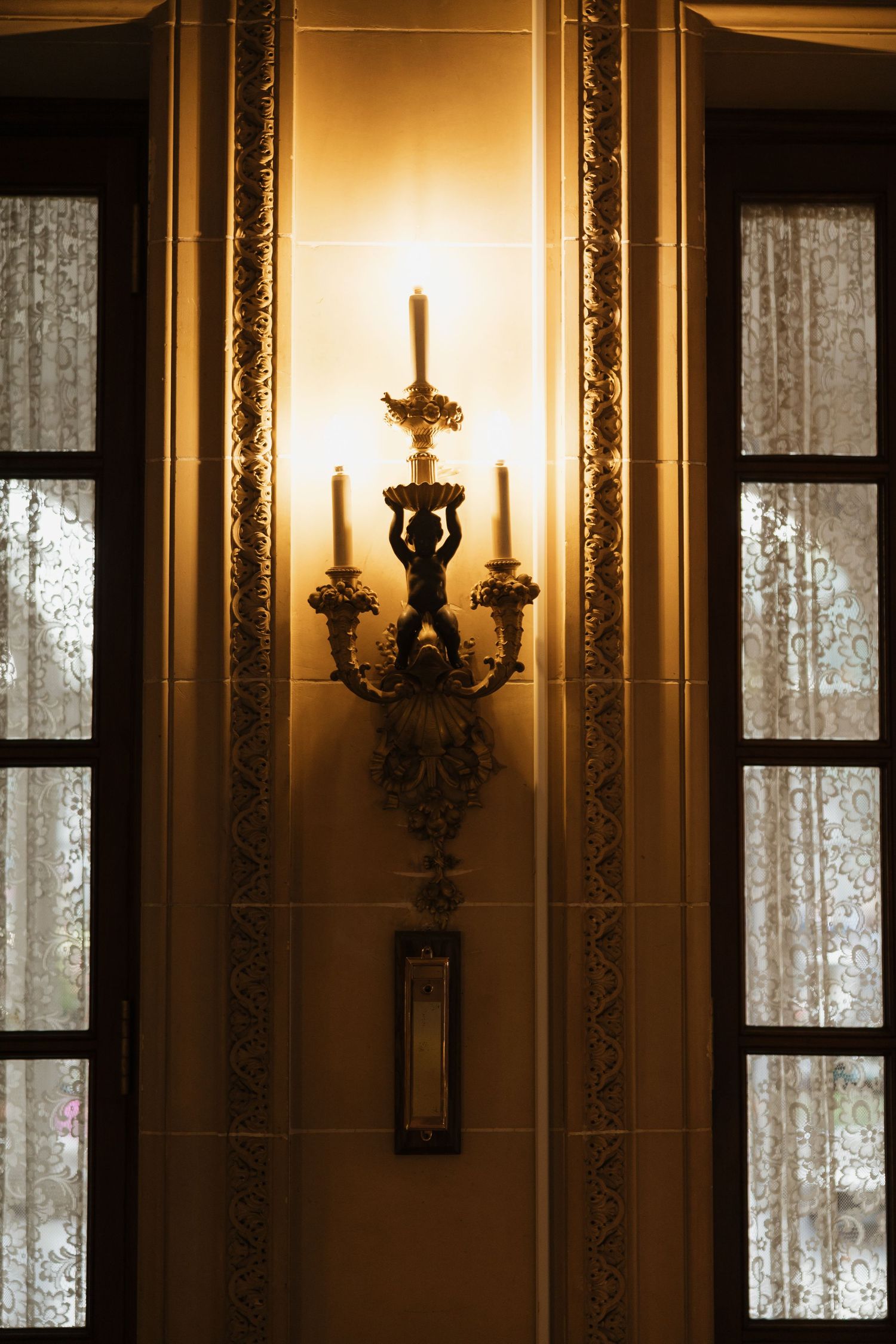 Ornate golden candelabra wall sconce with figurine detail illuminated between two windows with lace curtains.