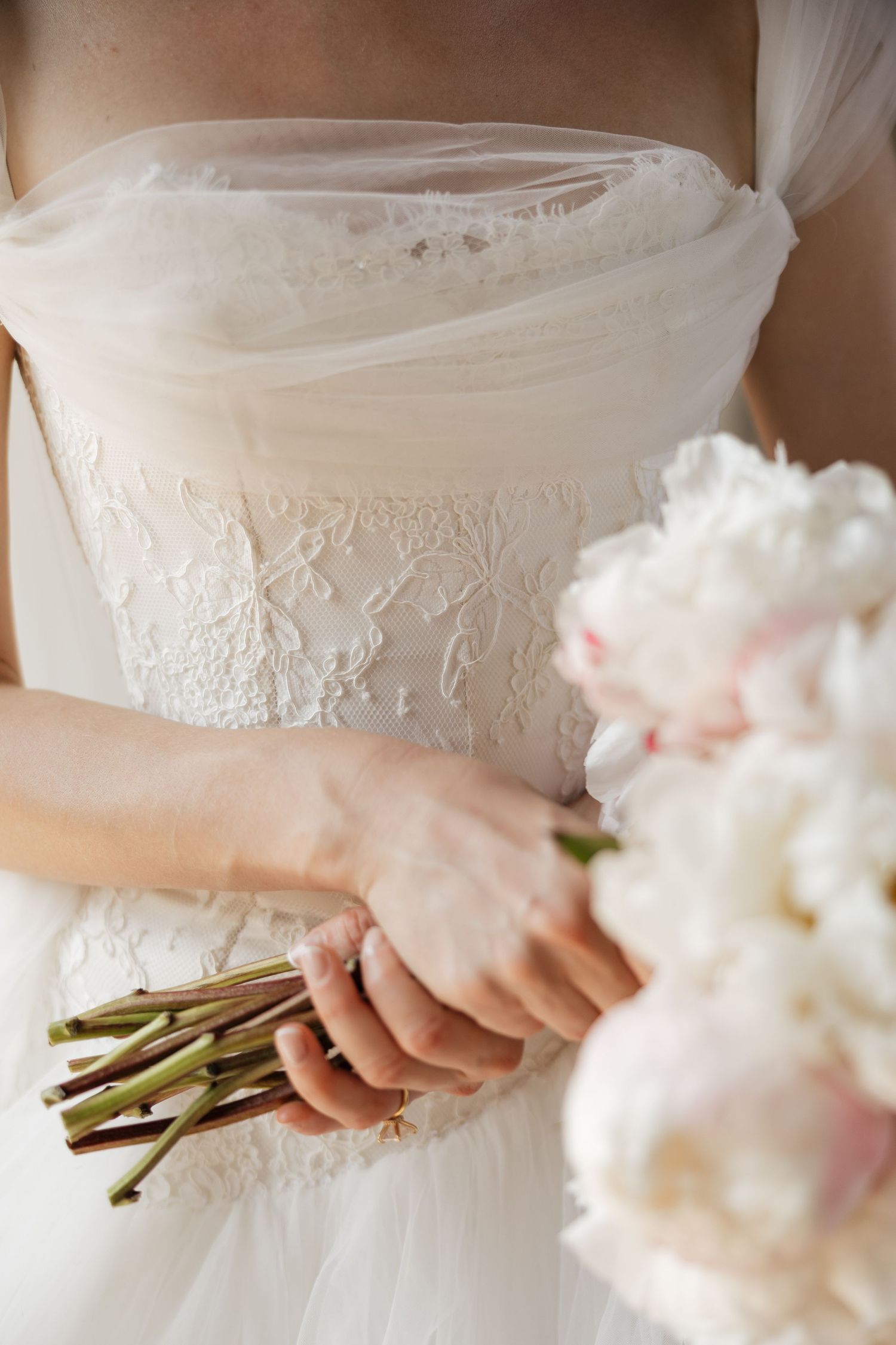 Bride in white lace strapless gown holding elegant bouquet of white and pink peonies at wedding ceremony.