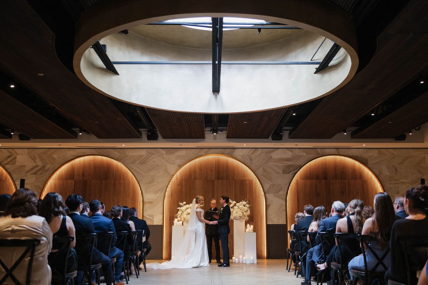 Elegant wedding ceremony in a modern venue with arched wood panels, circular skylight, and guests seated in rows.