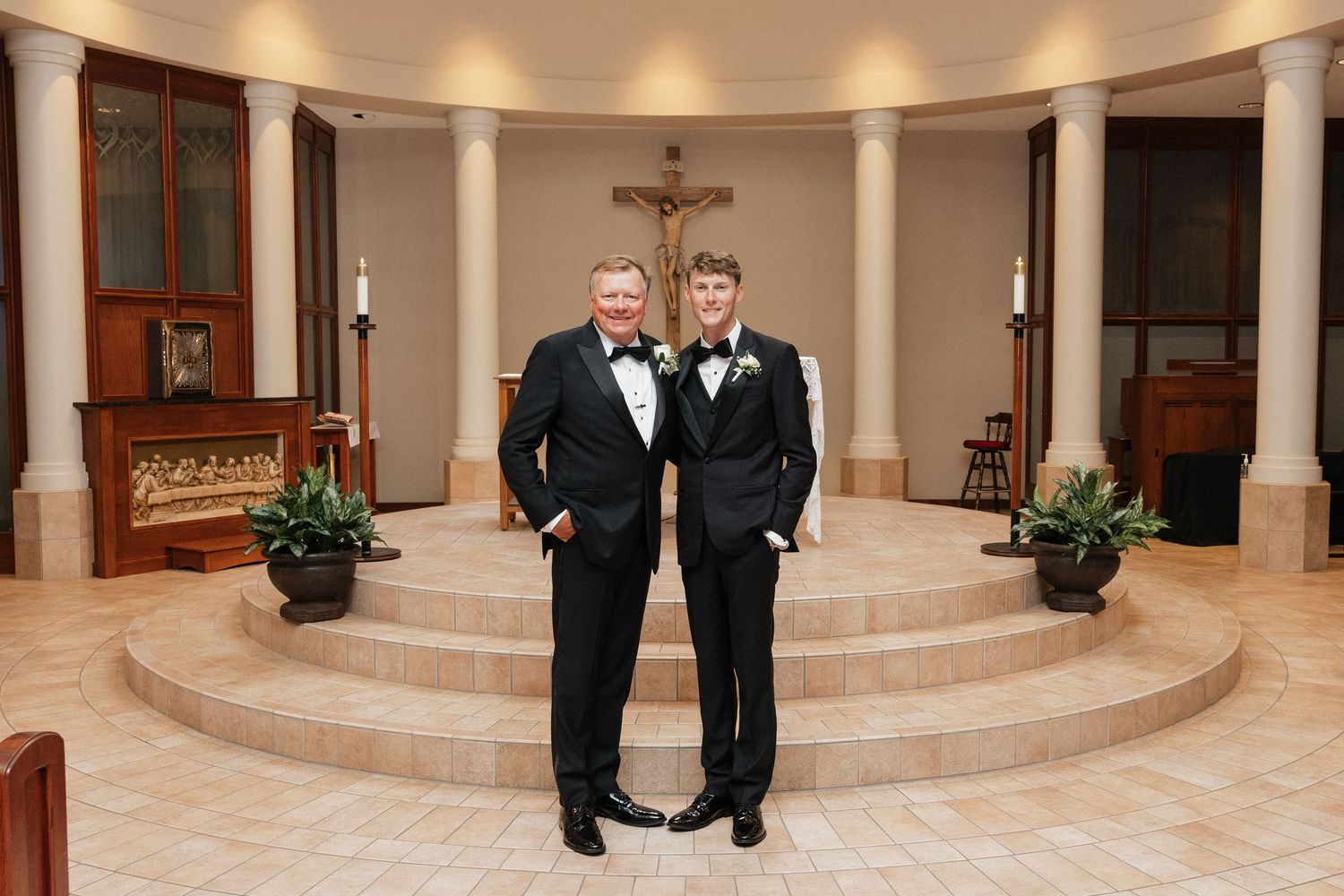 Two men in tuxedos pose in a church with a crucifix and altar backdrop at a wedding ceremony.