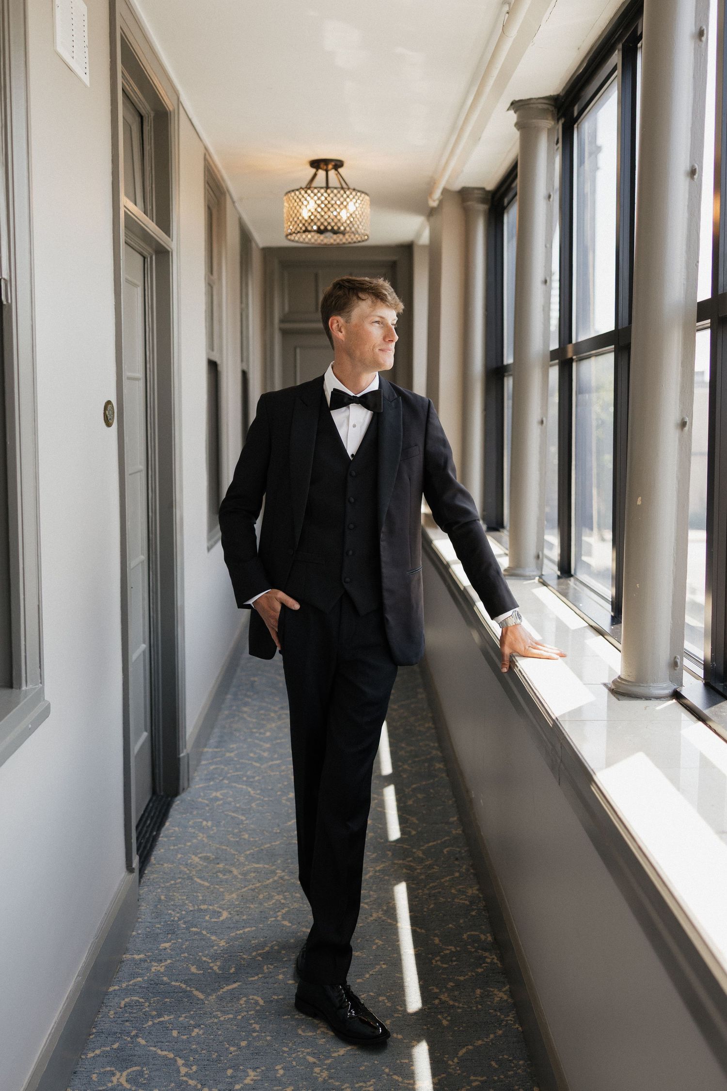 Elegant groom in black tuxedo with bow tie standing in a hallway, gazing out large windows.