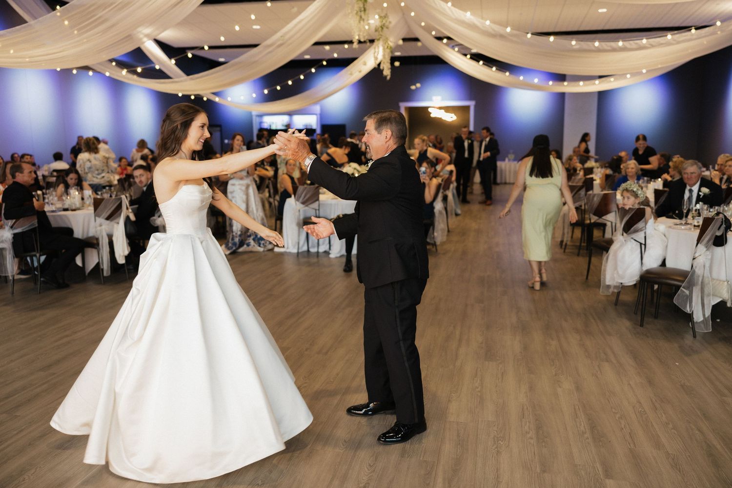 Bride and groom share their first dance at elegant wedding reception with draped ceiling lights and guests seated at tables.