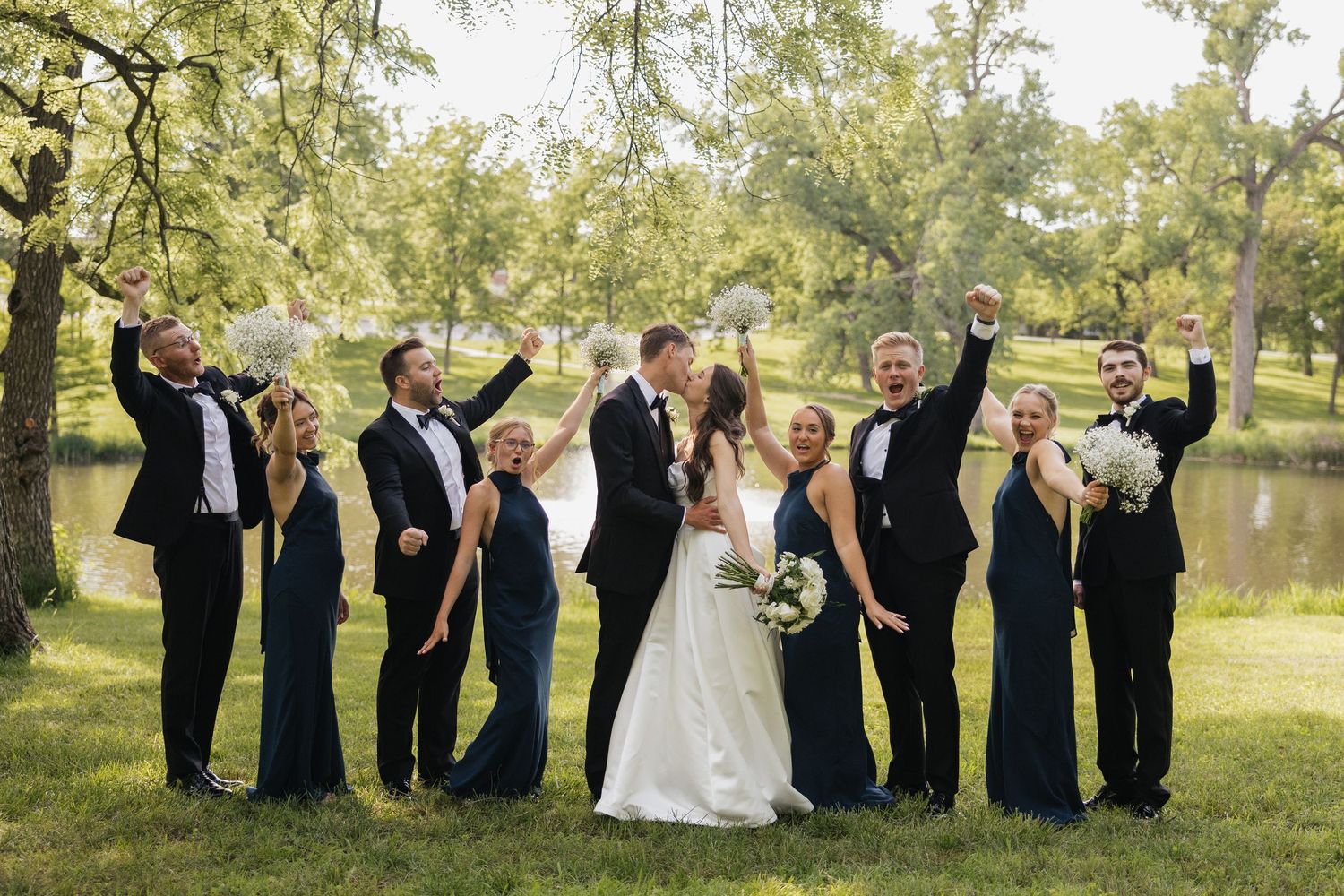 Wedding party in navy attire celebrating outdoors, bride and groom kissing while guests cheer near a pond.
