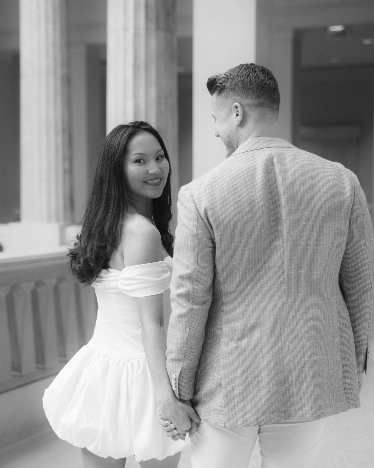 Black and white romantic photo of smiling woman in white dress holding hands with man in blazer near classical columns.