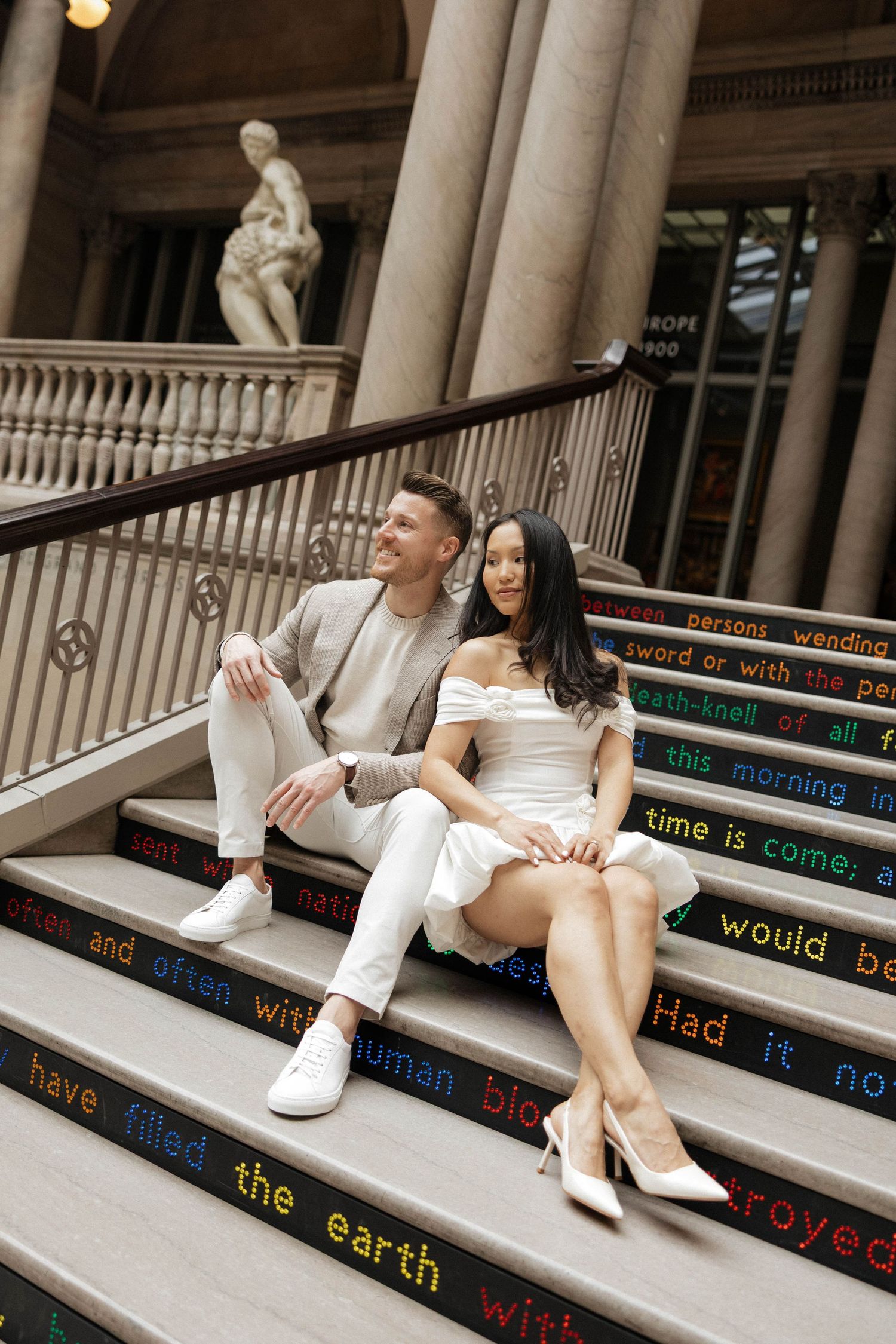 Bride and groom sitting on decorated steps with colorful text, posing beside classical columns and statue.