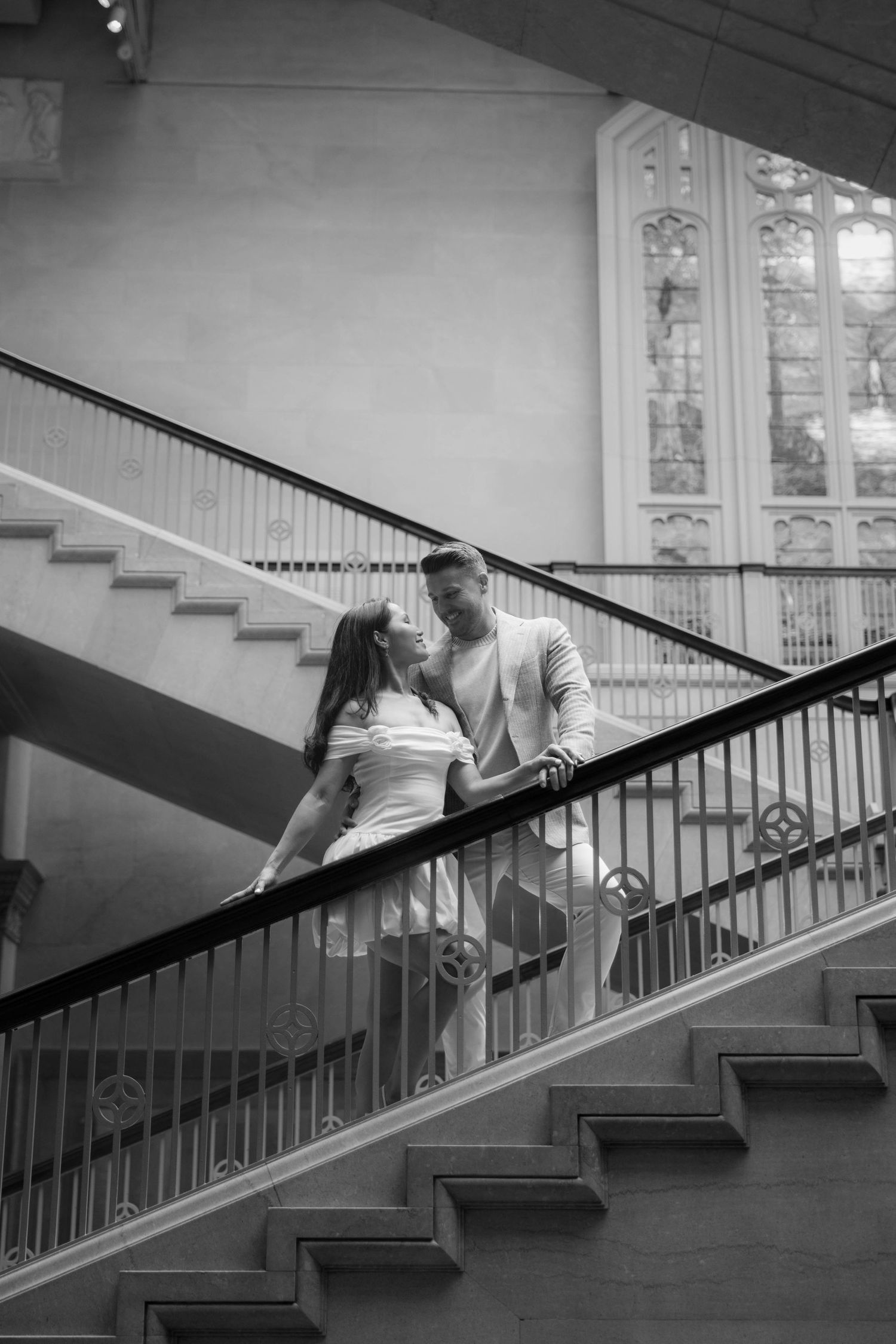 Black and white photo of romantic couple embracing on grand staircase with stained glass window.