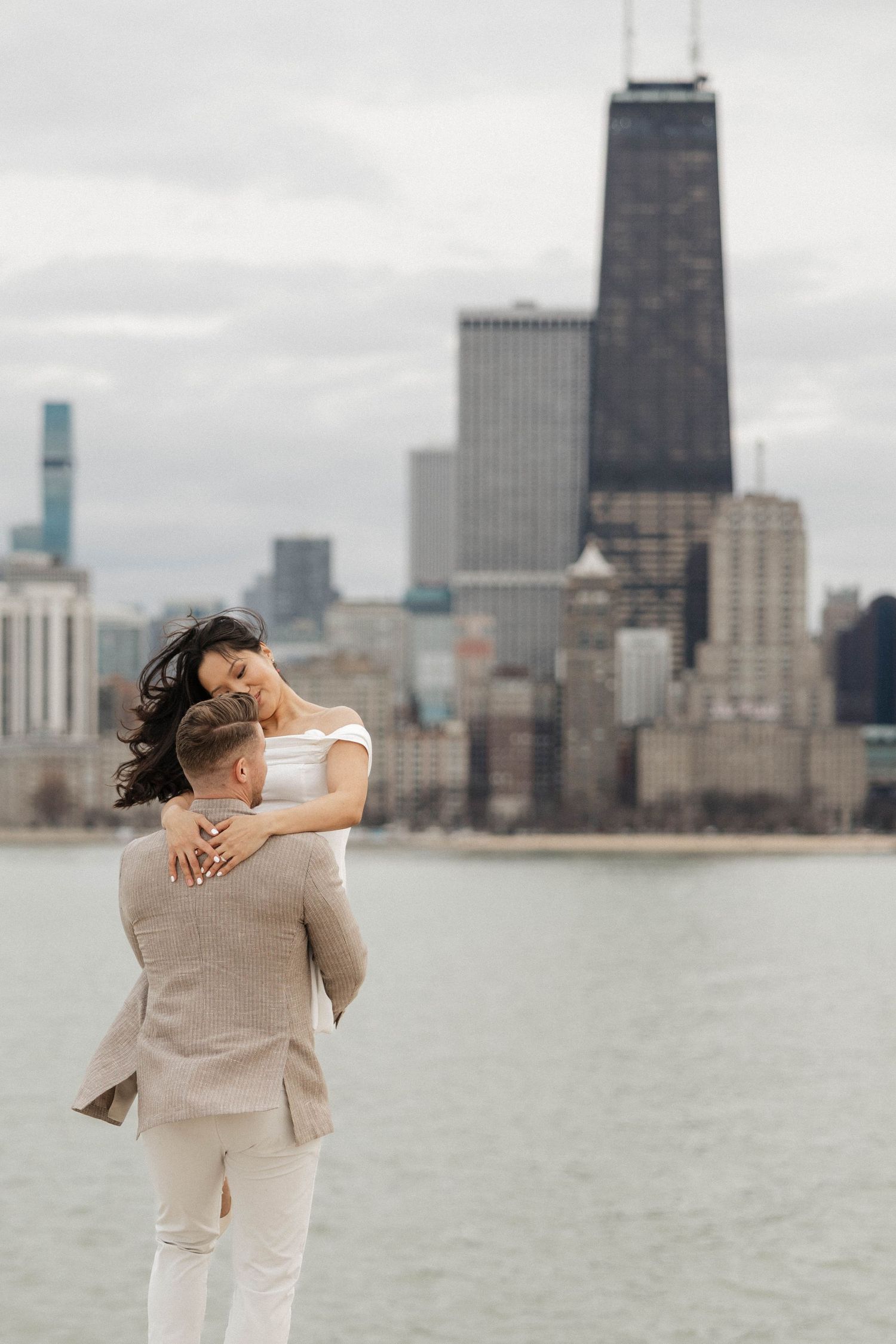 Couple embracing on Chicago waterfront with dramatic skyline featuring John Hancock Center in background.