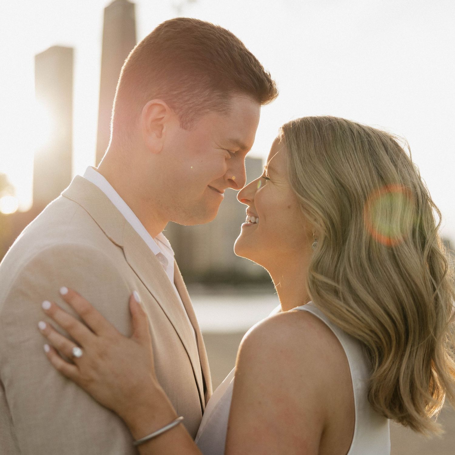 A couple shares a romantic moment at sunset, the man in a beige suit and woman in a sleeveless dress nearly kissing.
