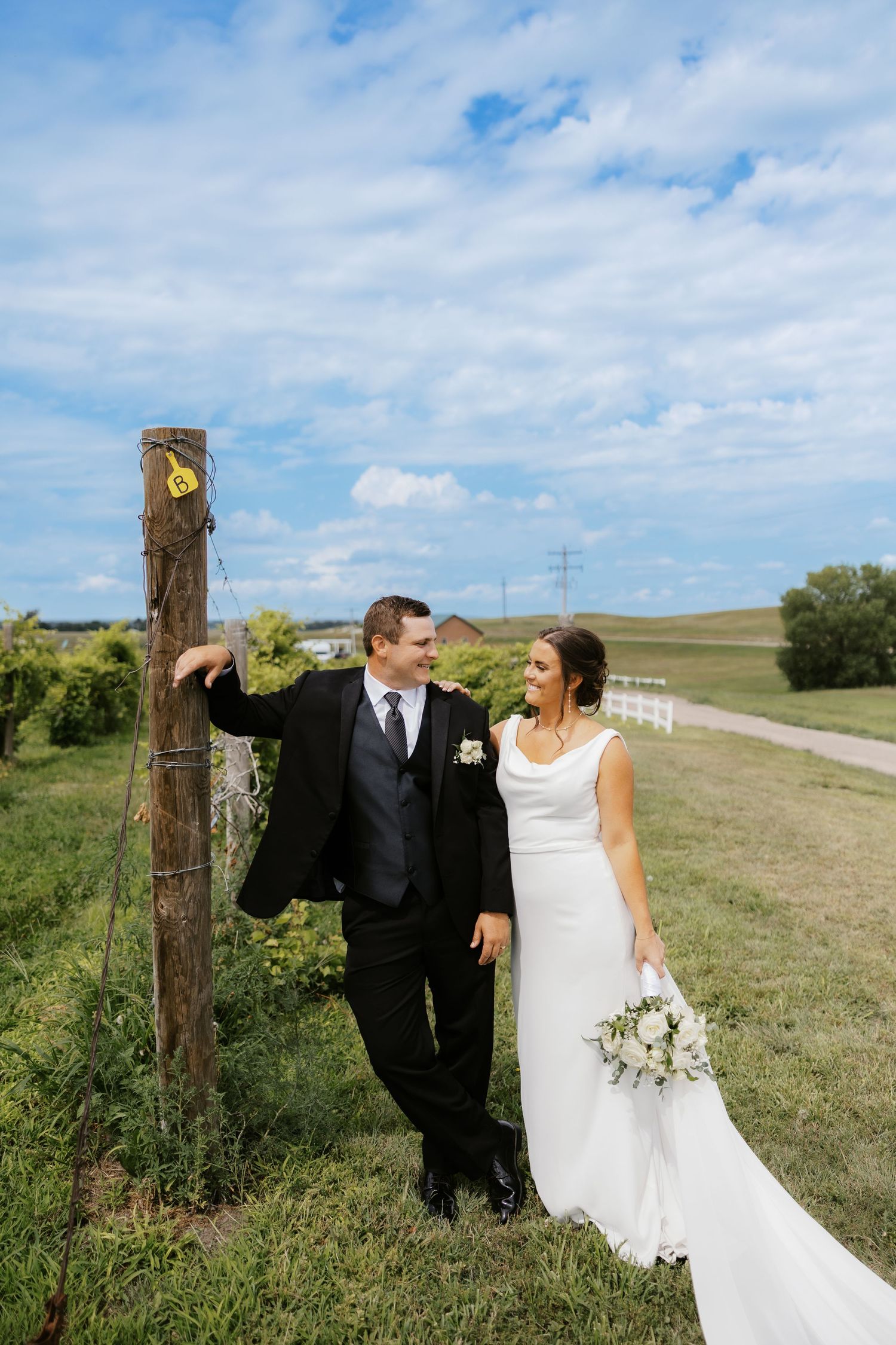 A couple in wedding attire poses together in a scenic rural field with rolling hills and blue skies.