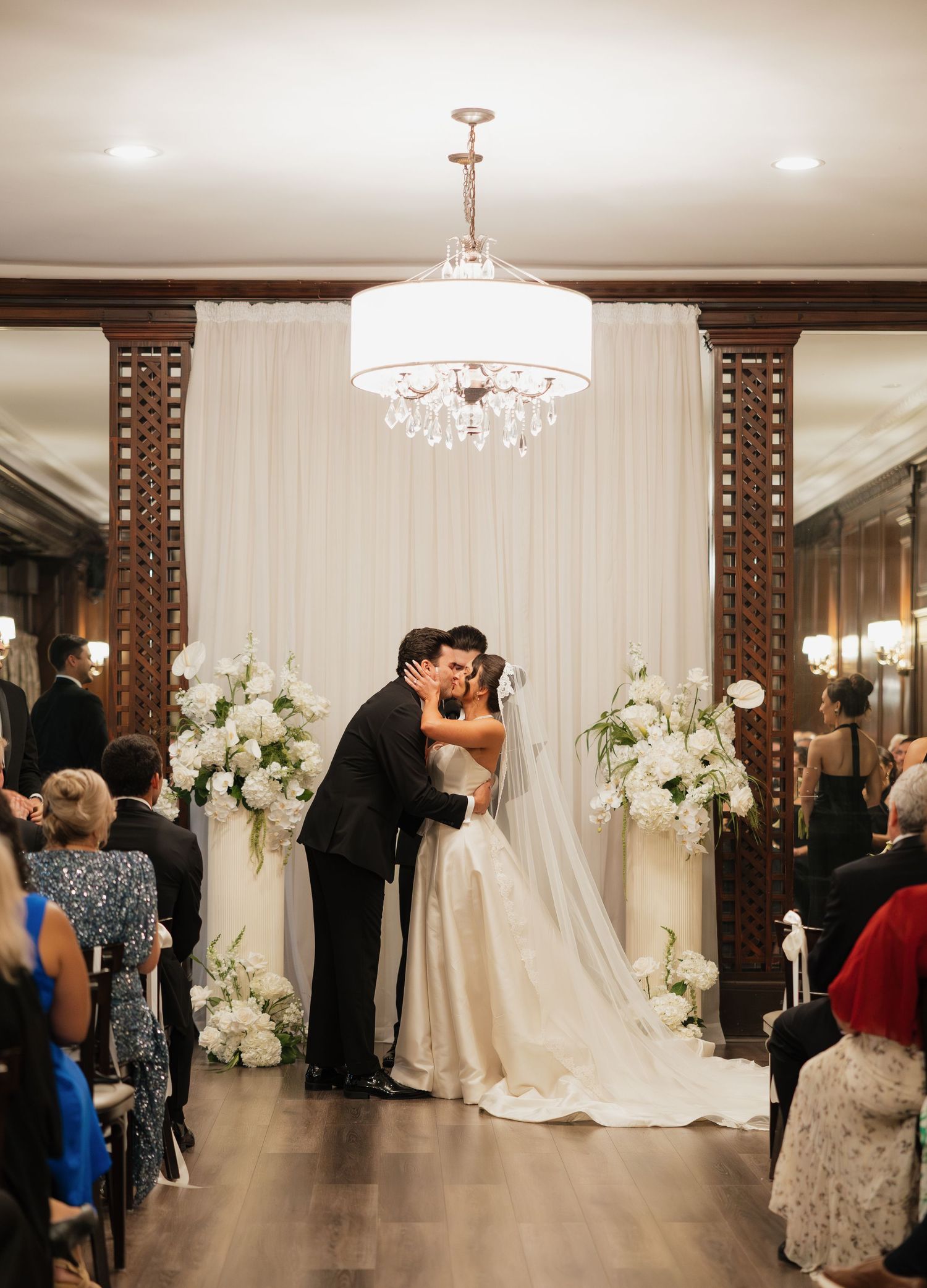 Bride and groom share first kiss at elegant indoor wedding ceremony with white floral arrangements and crystal chandelier.