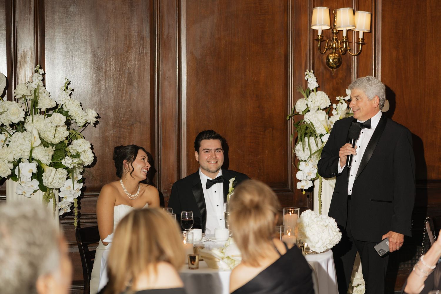 Elegant wedding reception toast in wood-paneled ballroom with white floral arrangements as older man speaks to smiling bride and groom.