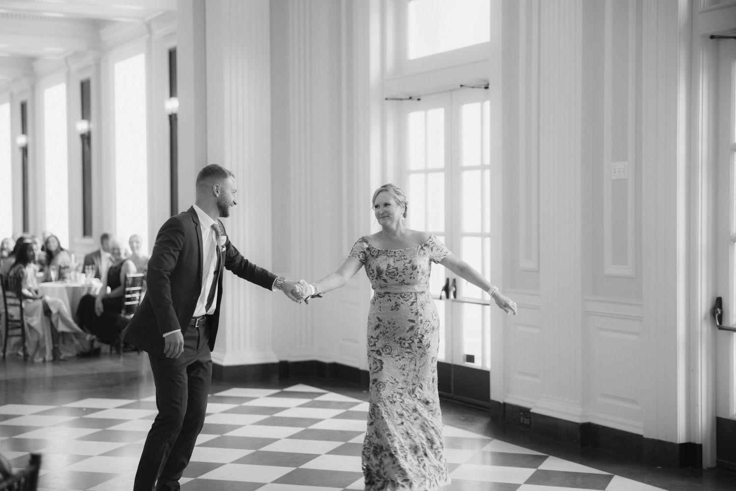 Two people dance gracefully in an elegant white ballroom with tall windows and columns.
