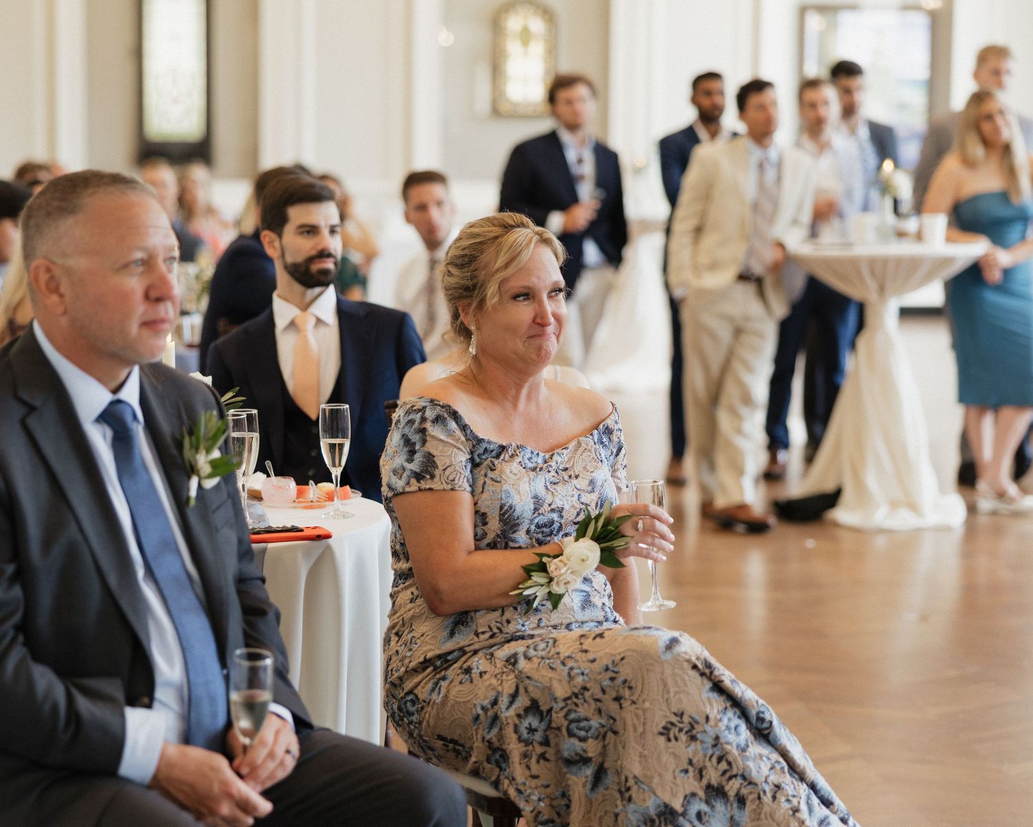 Wedding guests in formal attire gather at an elegant indoor reception venue with champagne glasses.
