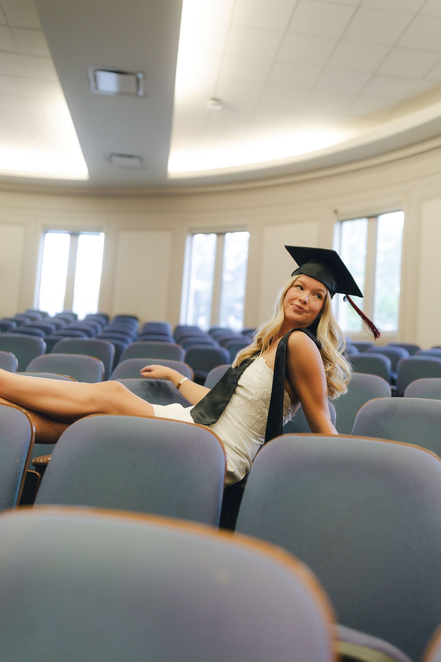 Graduate in cap and gown poses playfully across auditorium chairs in empty lecture hall.