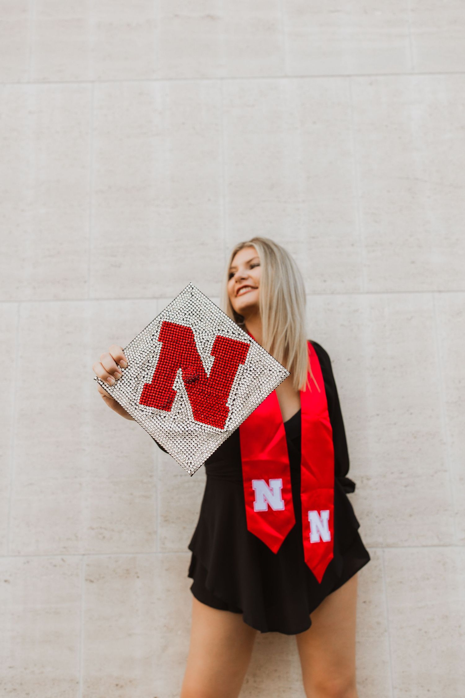 Graduate displays decorated graduation cap and red stole with N logo against white wall in celebratory pose.