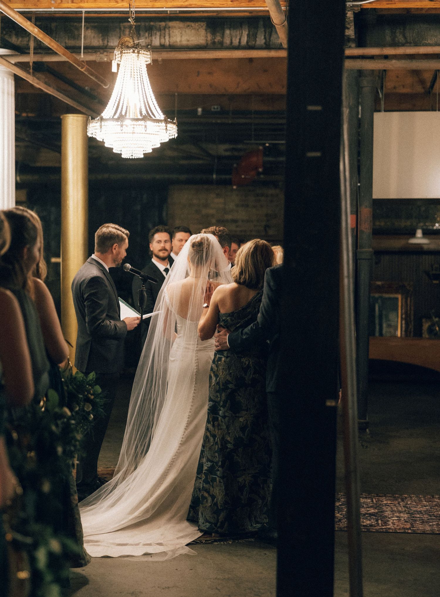 A romantic wedding couple shares an intimate dance moment under a crystal chandelier in an industrial-style venue.