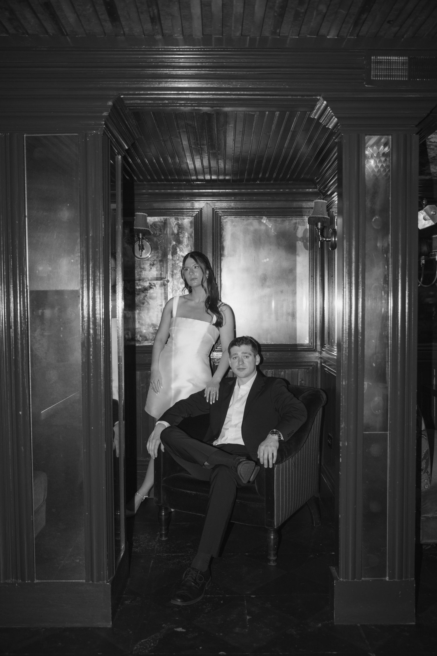 A stylish couple poses in an elegant vintage bar interior with dark wood paneling and moody lighting.