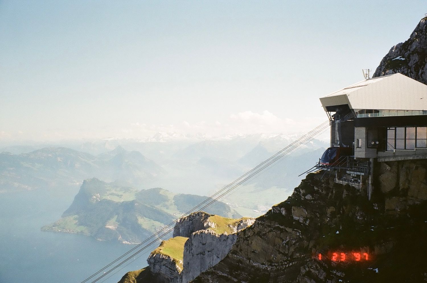 Scenic mountain landscape with a modern building perched on a snowy cliff overlooking majestic snow-capped peaks at sunset.