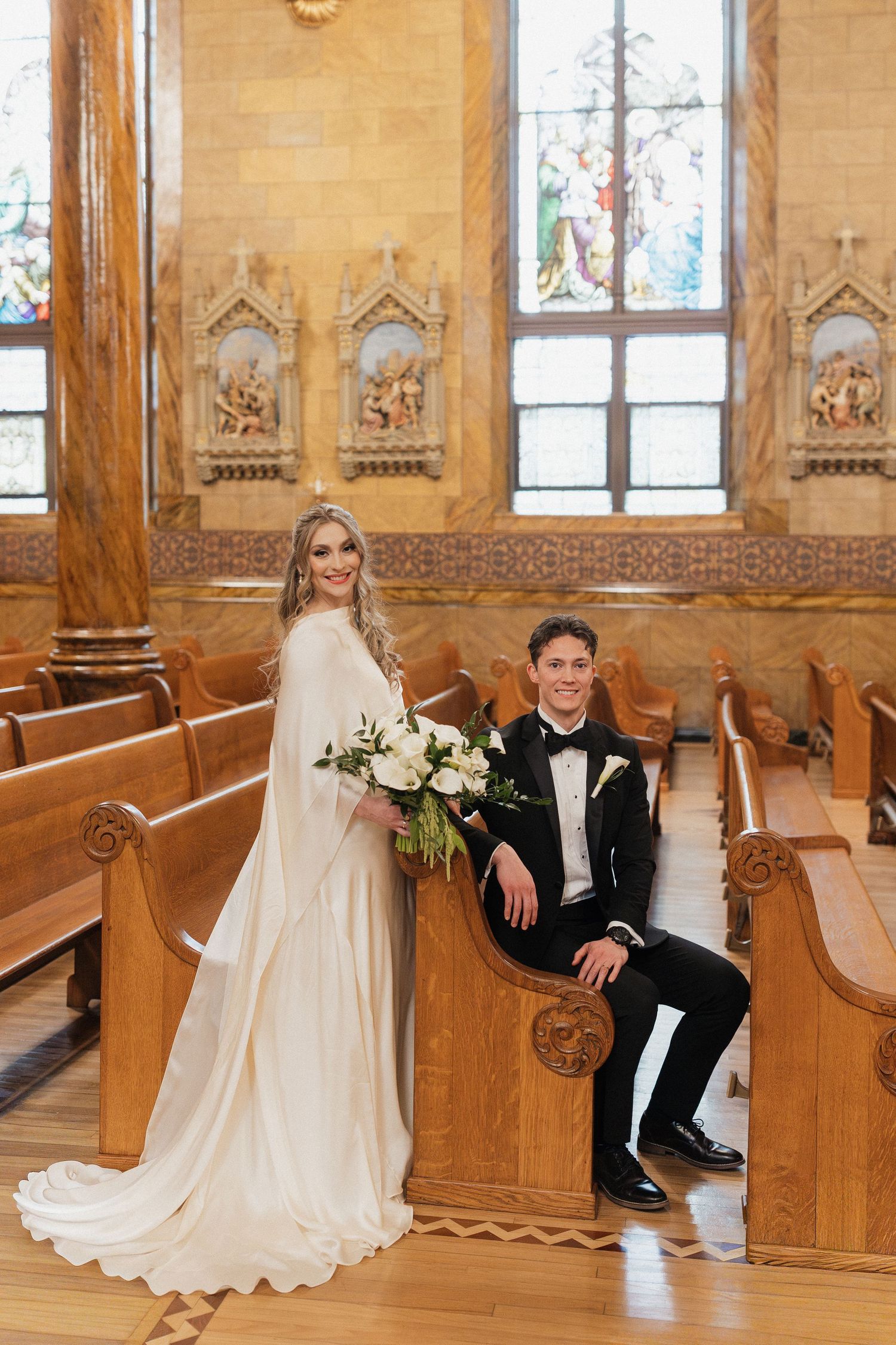Wedding couple sitting in ornate wooden church pews with stained glass windows and decorative carvings.