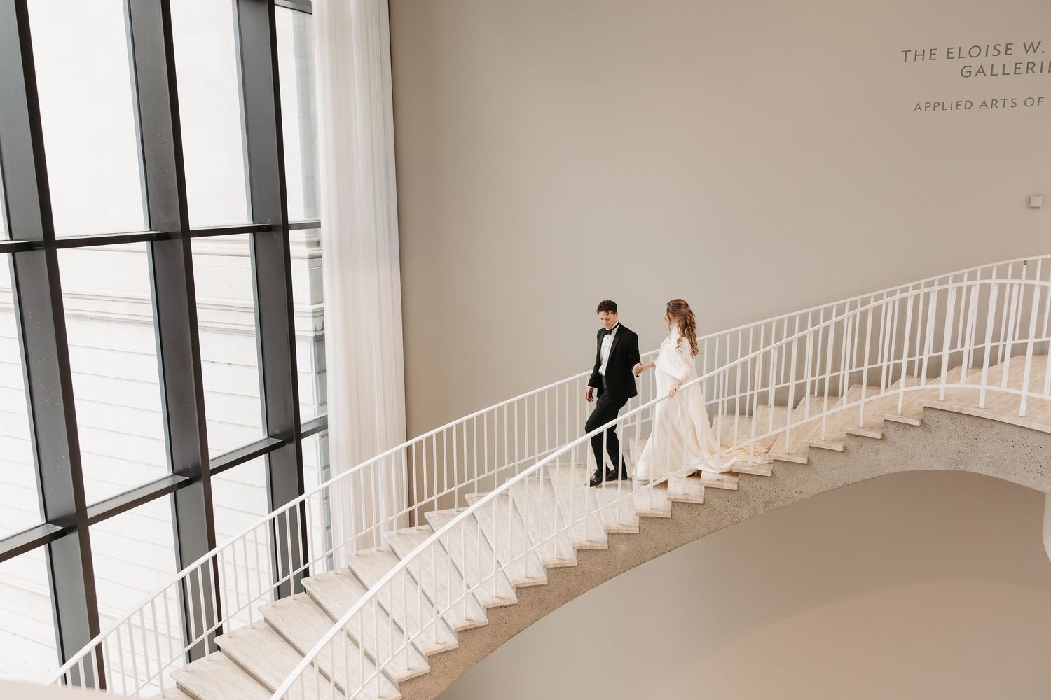 Couple in wedding attire walking down elegant white spiral staircase in modern art gallery with tall windows.