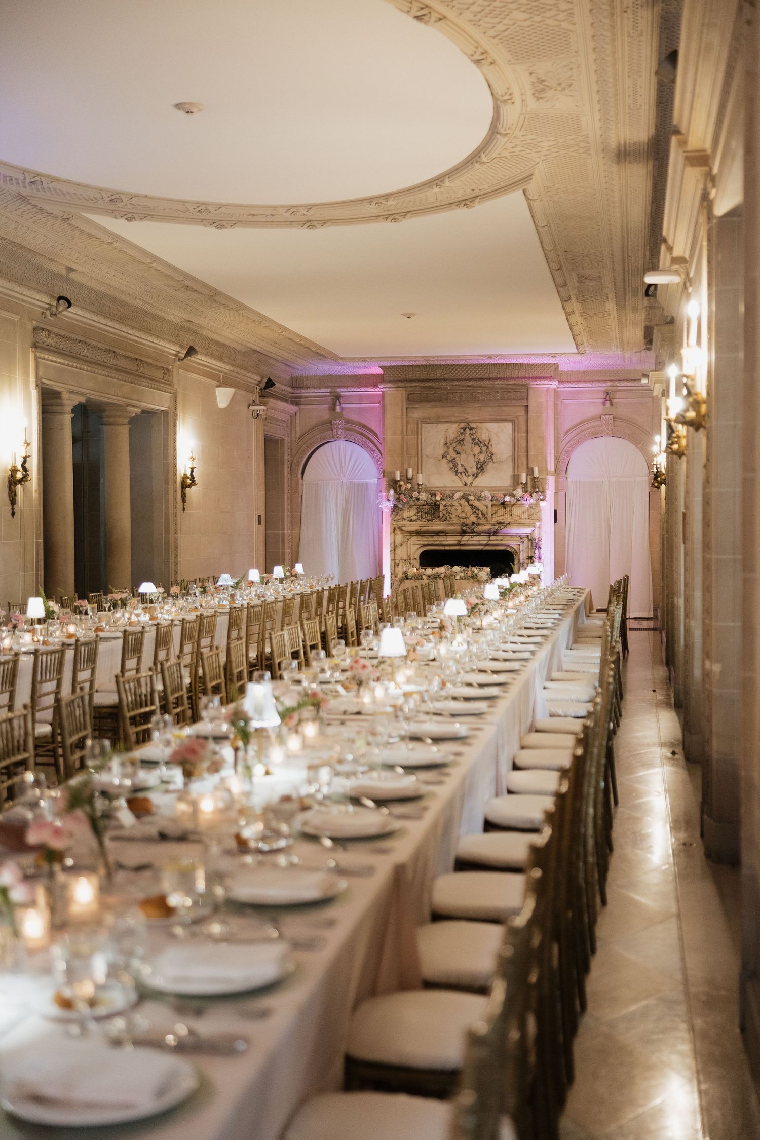 Elegant wedding reception hall with long banquet tables, gold chiavari chairs, candlelight, and ornate architectural details.
