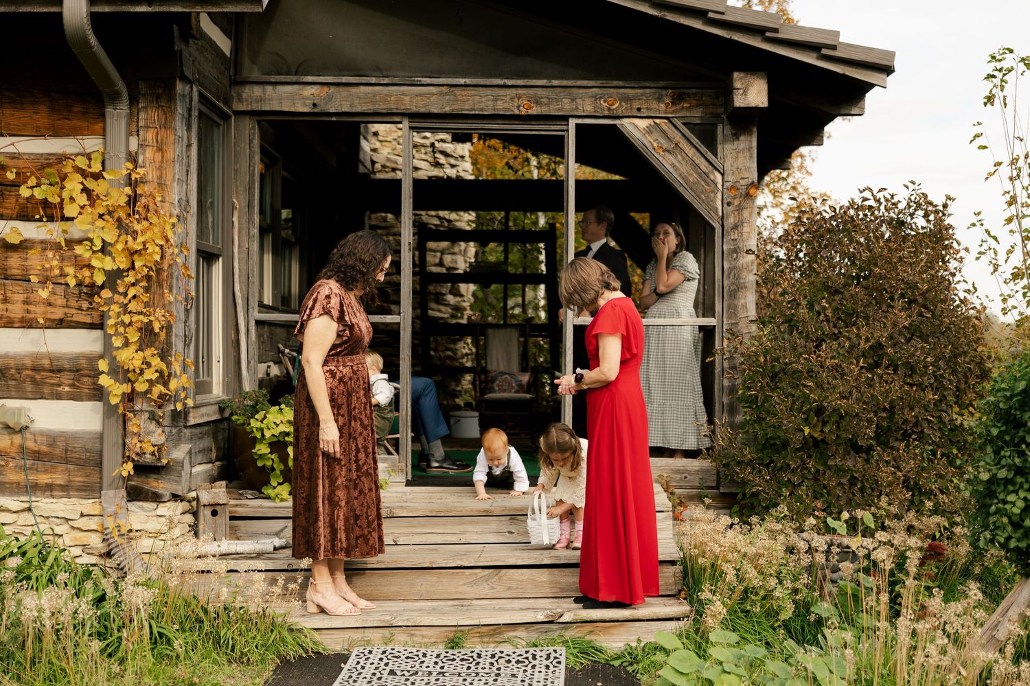 Two women in dresses stand on rustic wooden steps greeting toddlers outside a weathered cabin surrounded by lush autumn garden greenery.