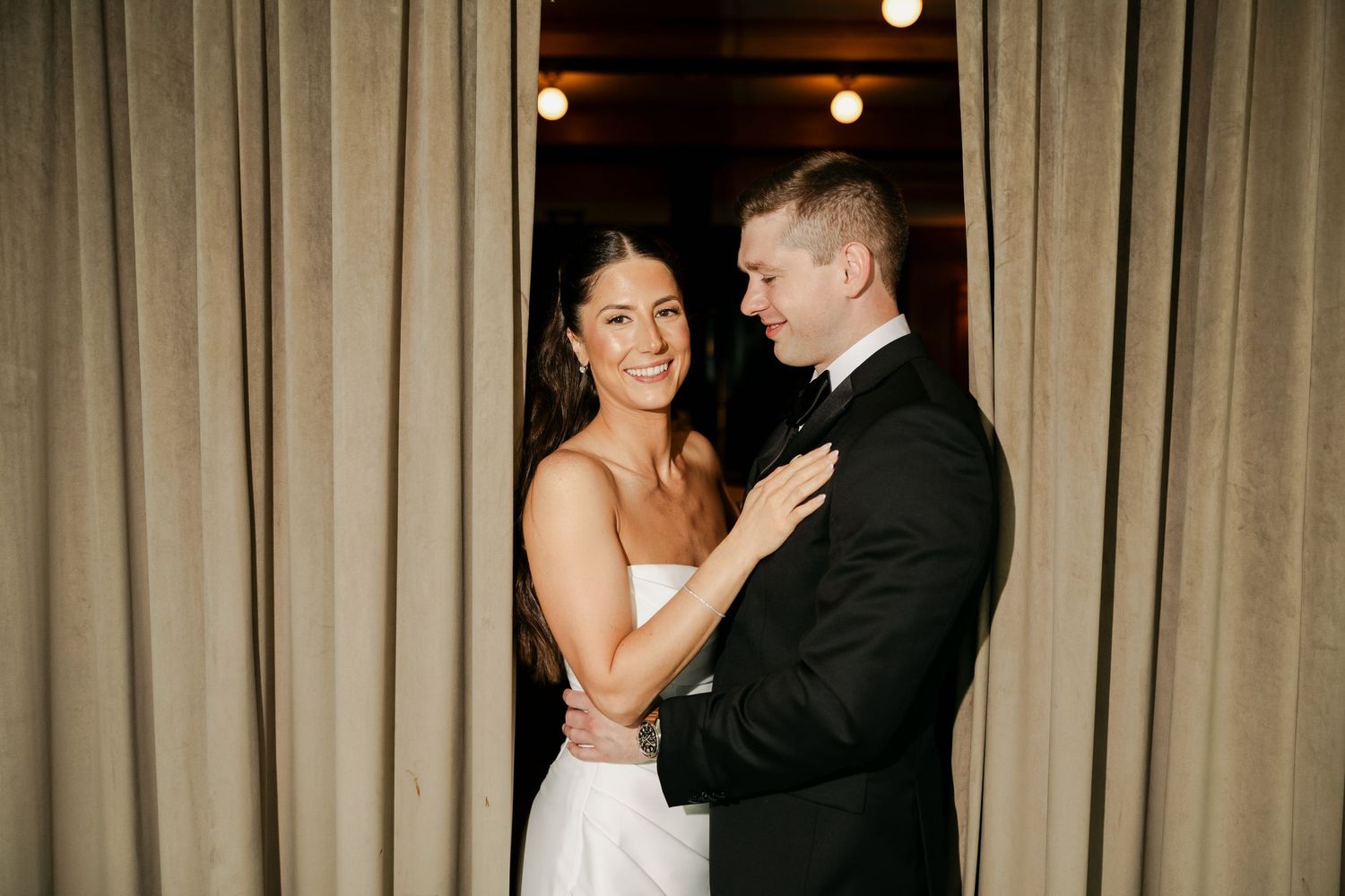 Smiling bride in a white strapless dress embraces her groom in a dark suit beside draped curtains and warm ambient lighting.