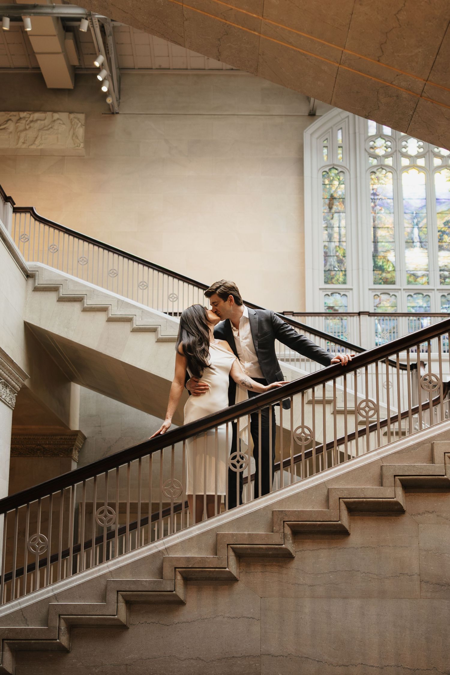 Couple embracing on a grand marble staircase with ornate railings beneath a large stained glass window during an engagement photo session.