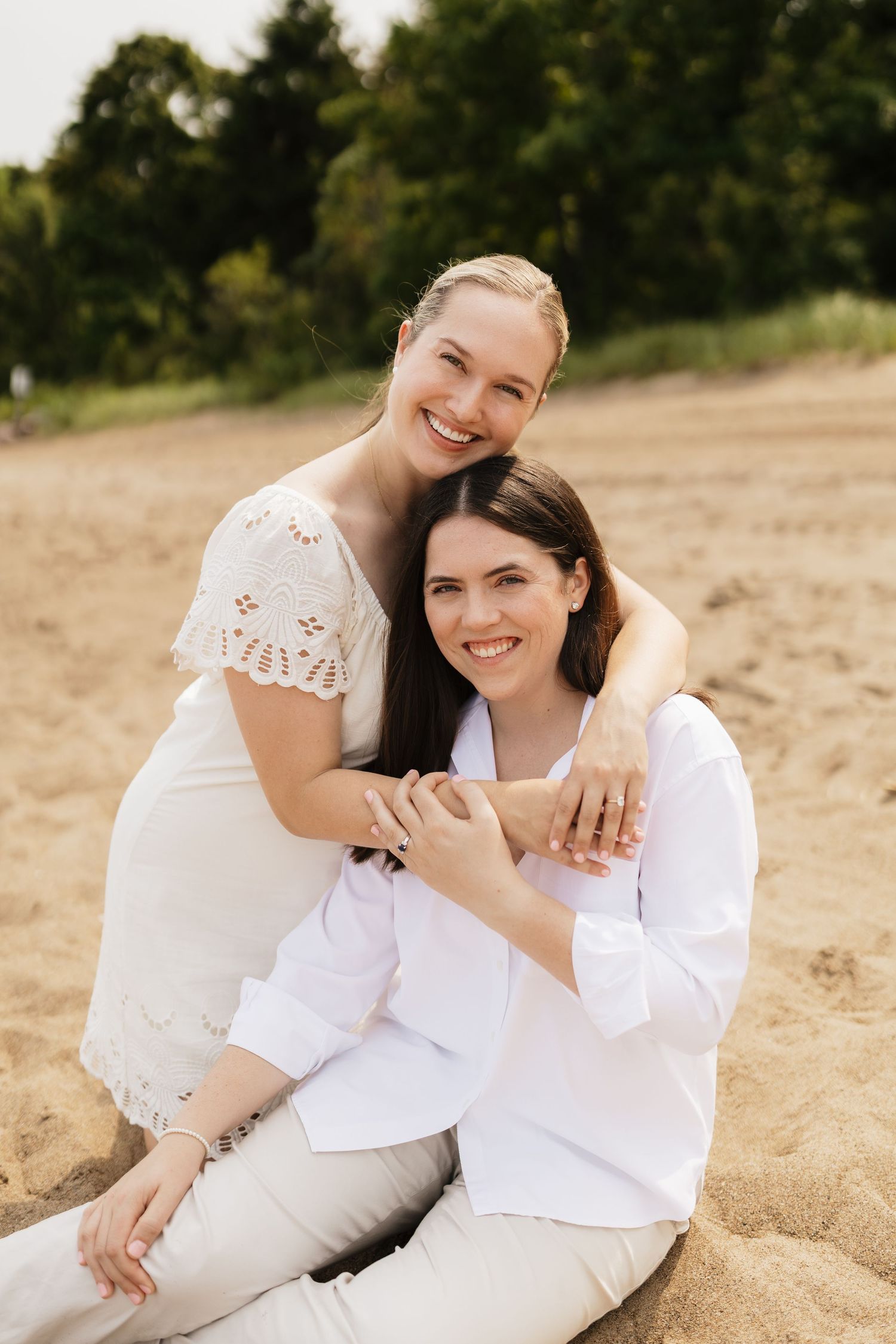 Two smiling women in white outfits sitting together on a sandy beach, sharing a warm embrace.