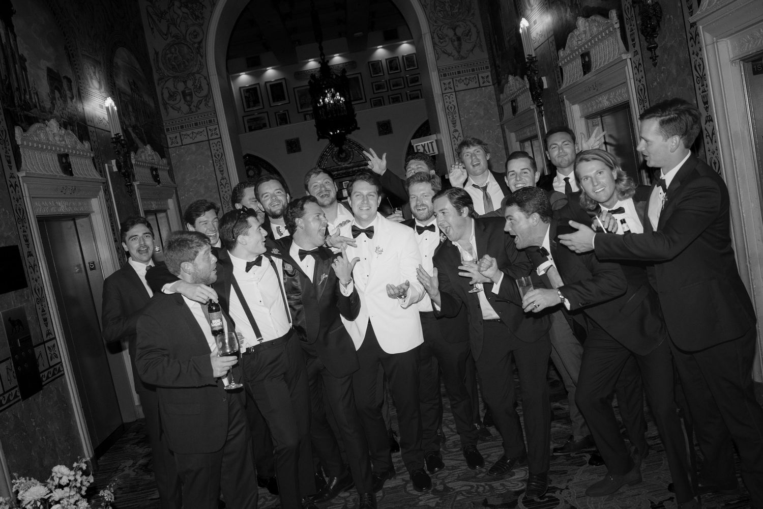 Black and white photo of groom with groomsmen in tuxedos celebrating at elegant wedding reception venue.