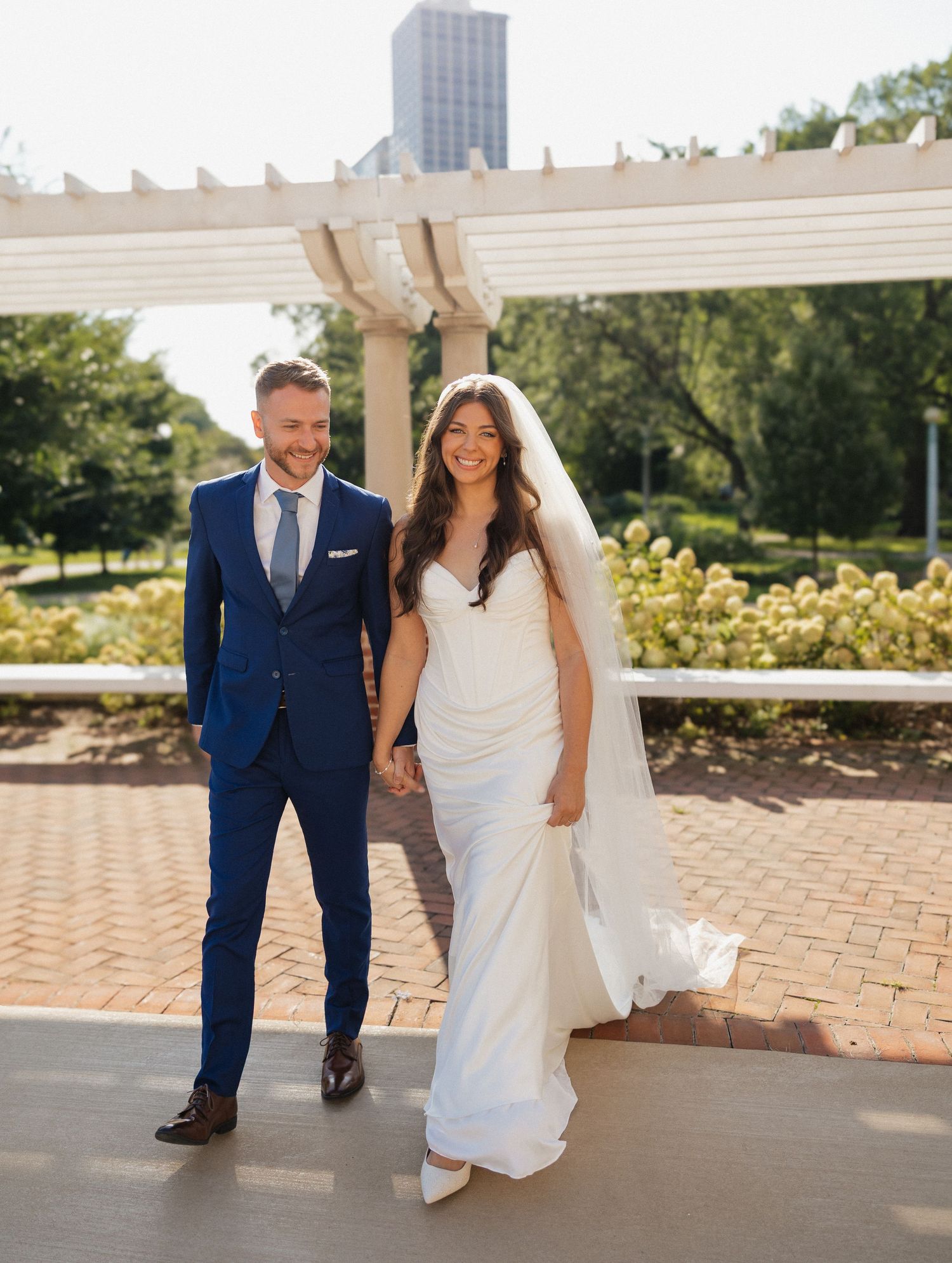A bride and groom walk hand in hand along a brick pathway on their wedding day in a sunny outdoor setting.
