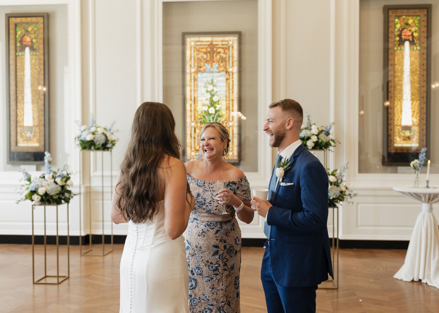 Guests gathered in an elegant ballroom with ornate white walls, stained glass windows, and floral arrangements.