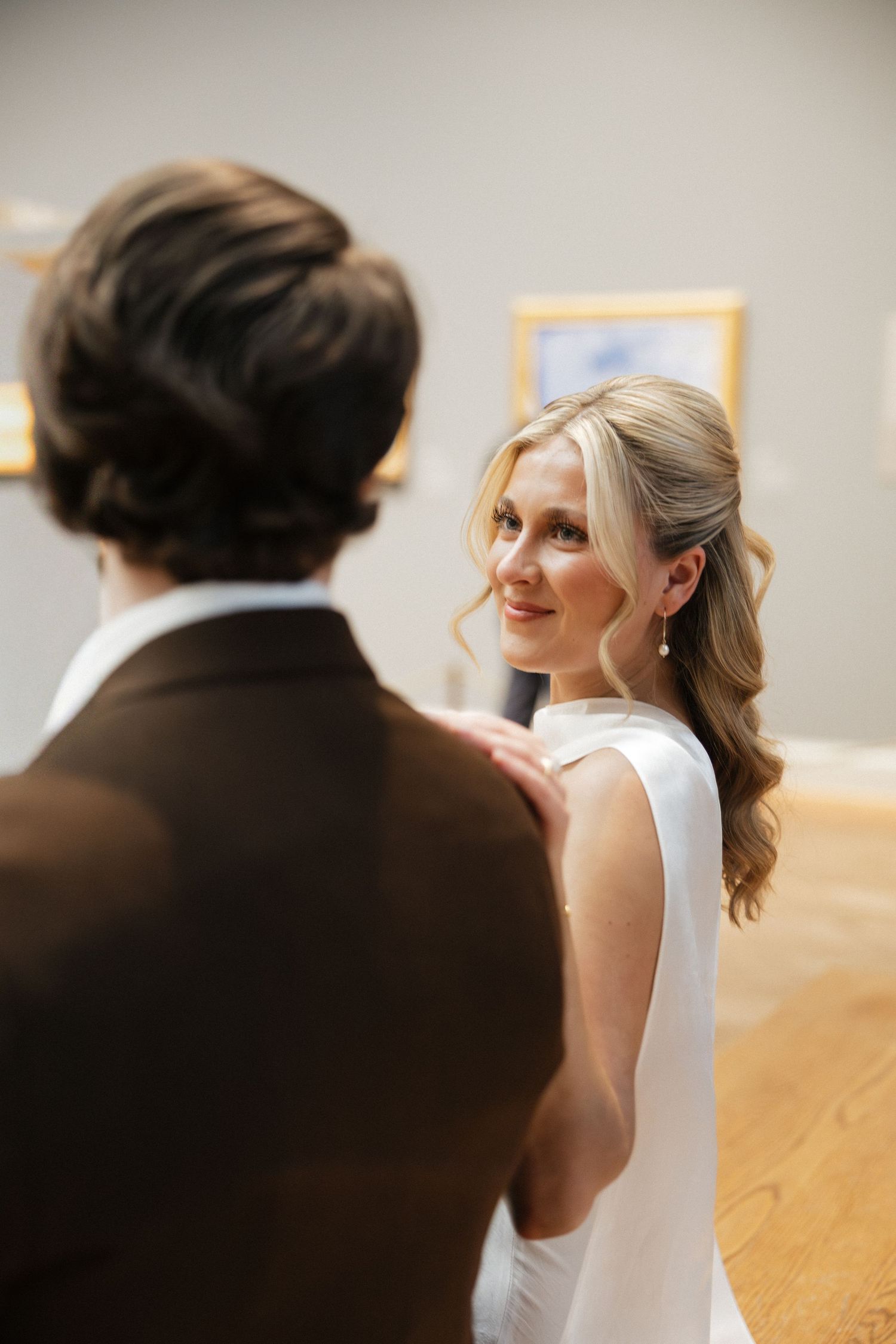 Bride in white dress smiles at groom in brown suit during wedding ceremony in elegant gallery venue.