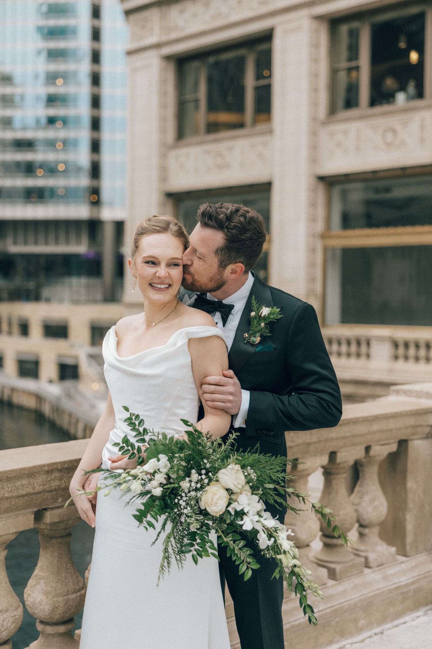 A bride and groom share an intimate moment on stone steps of a historic building with a cascading white floral bouquet.