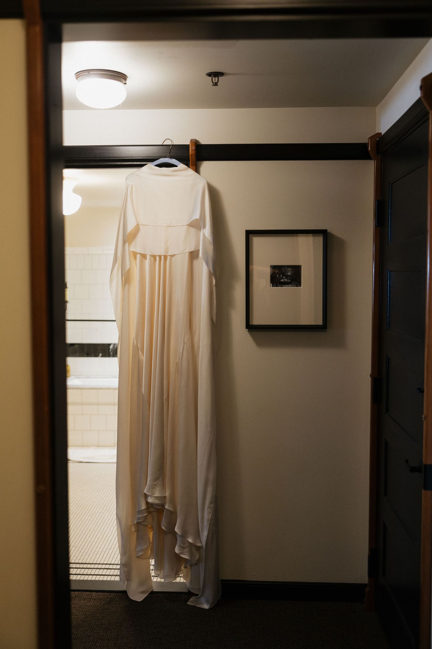 A white wedding dress hangs elegantly in a dimly lit hallway against a cream-colored wall.